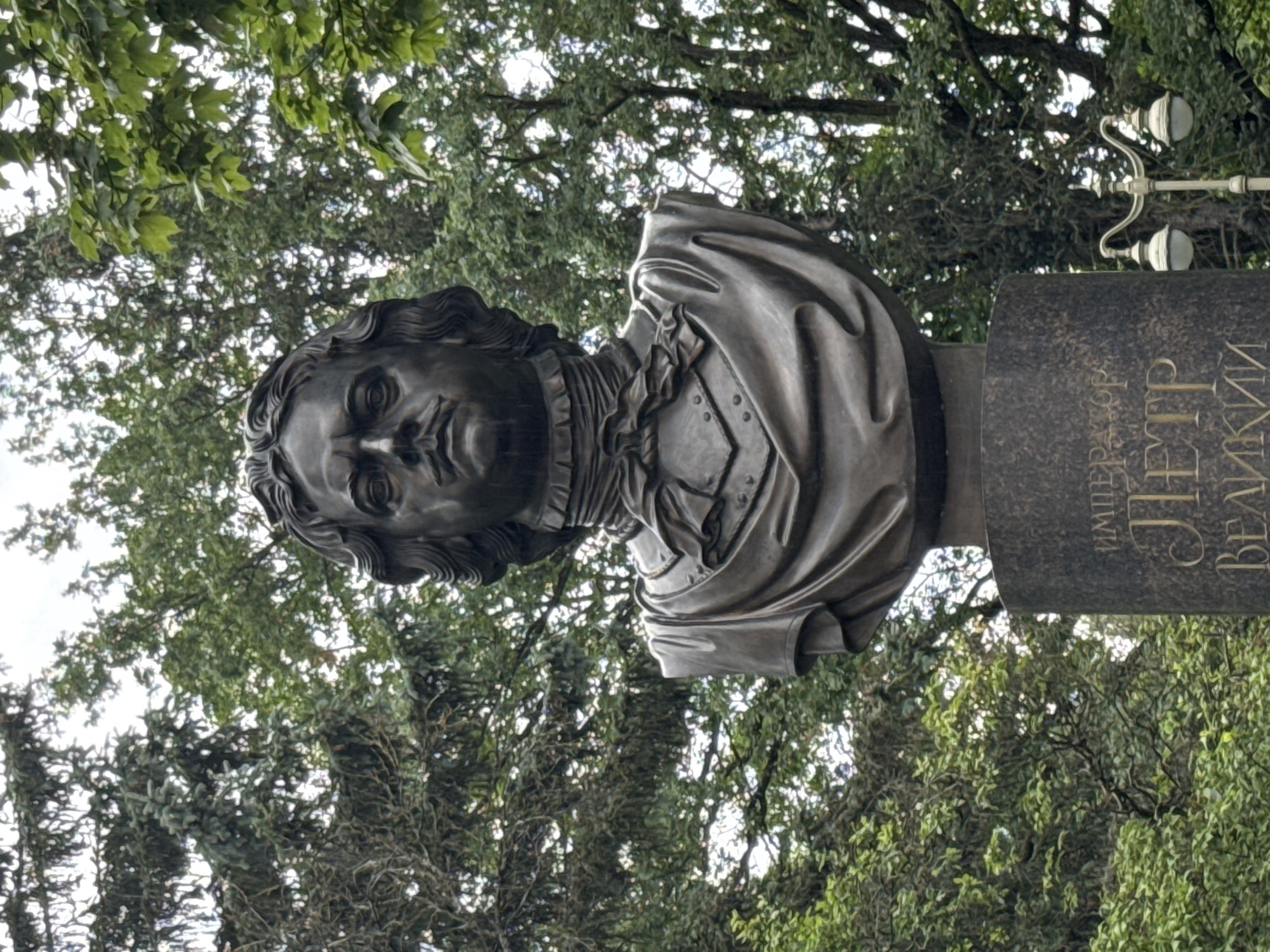 A bronze bust of a man with a stern expression, a mustache, and curly hair is positioned on a dark stone pedestal. The bust depicts him wearing a ruffled collar and a chest plate with decorative detailing. The pedestal has Cyrillic inscriptions. Behind the bust, dense green trees and foliage form the background, with some evidence of rain or moisture on the statue. A lamppost with two lights is visible to the right of the pedestal. The scene appears to be outdoors, possibly in a park or public square. The inscription on the pedestal reads "ИМПЕРАТОР ПЕТР ВЕЛИКИЙ," which translates to "Emperor Peter the Great." This statue is located in Sinyavino, Russia.
