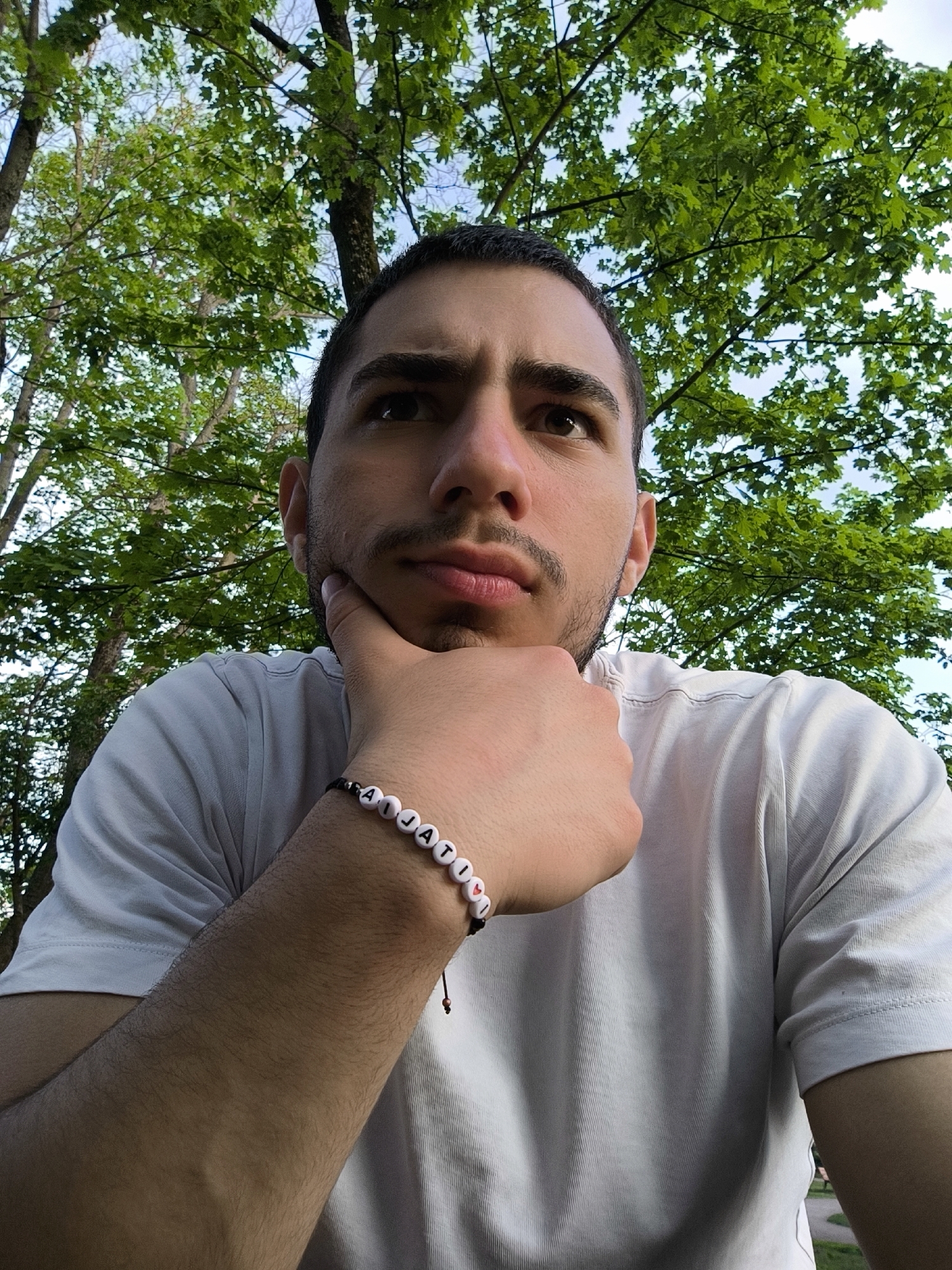 A young man with dark hair and a light complexion looks upward, with his chin resting on his hand. He is wearing a white t-shirt and a bracelet with the name "Aja Ti" spelled out in beads. The background shows lush green foliage, suggesting a park or wooded area. The man is outdoors, likely in a natural or semi-natural setting. While there is no indication of the location of the city of Strasbourg, France, the image does not rule out it being located in a park in that city.