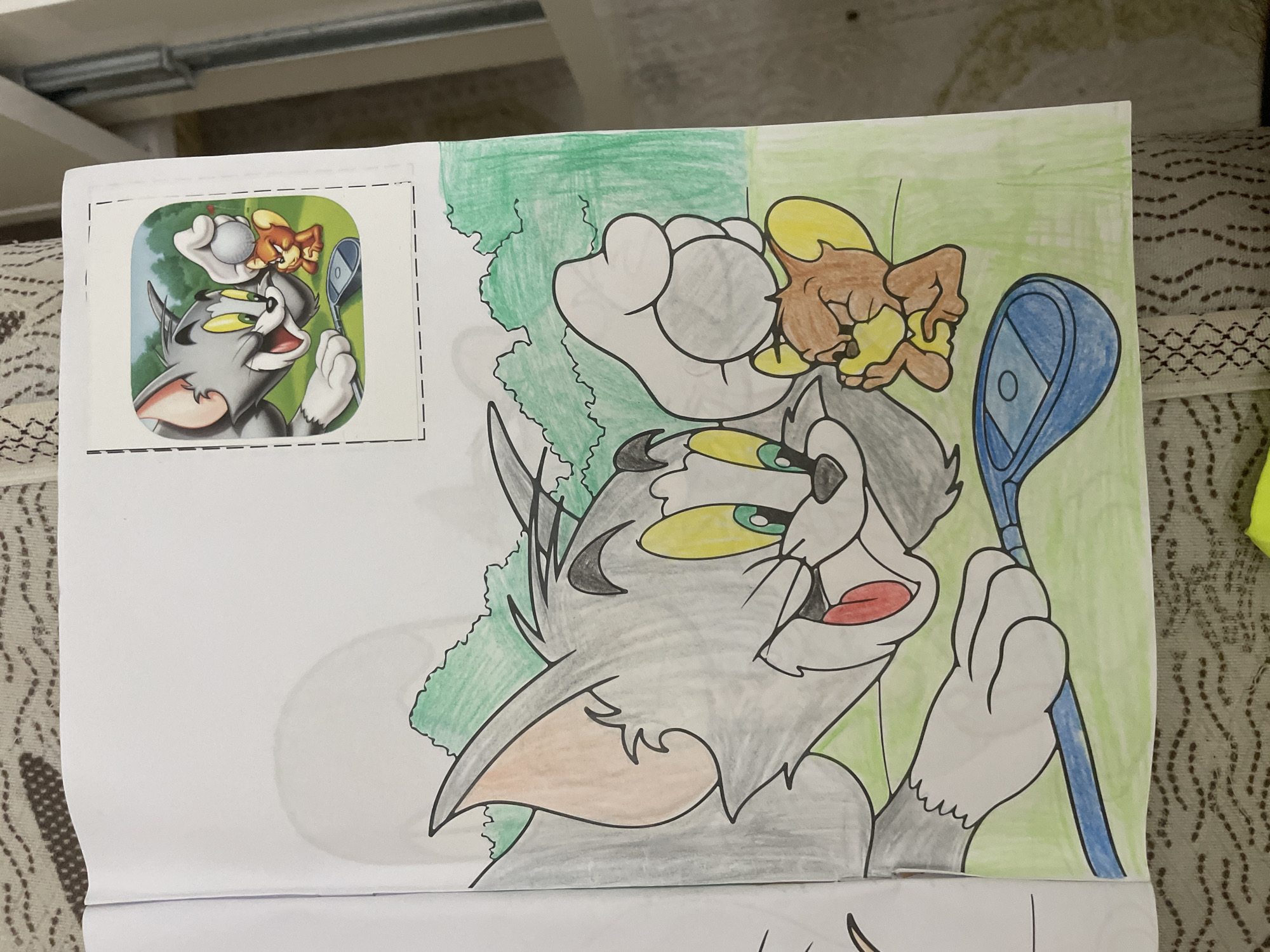 A hand-drawn coloring page featuring Tom and Jerry is displayed on a flat surface, likely indoors in Ihtiman, Bulgaria. The main subject is Tom, the gray cat, with Jerry, the brown mouse, positioned on his head/ear. Tom is depicted with wide, startled yellow eyes with green irises and an open mouth showing a red tongue, conveying surprise or shock. Jerry holds a small, light-colored object, implied to be a golf ball, and appears to be in the act of playing golf, using Tom's head as a tee. A blue golf club is held by Tom's right paw, but it's actually Jerry who is controlling the action.
The drawing is partially colored using pencils or crayons. Tom's fur is gray with darker shading and black outlines, and his nose is peach-colored. Jerry is colored brown with yellow accents. The background of the drawing is filled with two shades of green, suggesting an outdoor setting like a golf course or grassy area, with some areas left uncolored.
In the top-left corner of the image, there is a smaller, printed reference picture, framed by a dotted line, which served as the template for the coloring page. This reference image also shows Tom and Jerry in a golf scenario, with the number '0' visible on the golf club head. The overall scene captures a humorous, mischievous interaction between the classic cartoon characters. The image is brightly lit, showing the textures of the paper and the coloring.