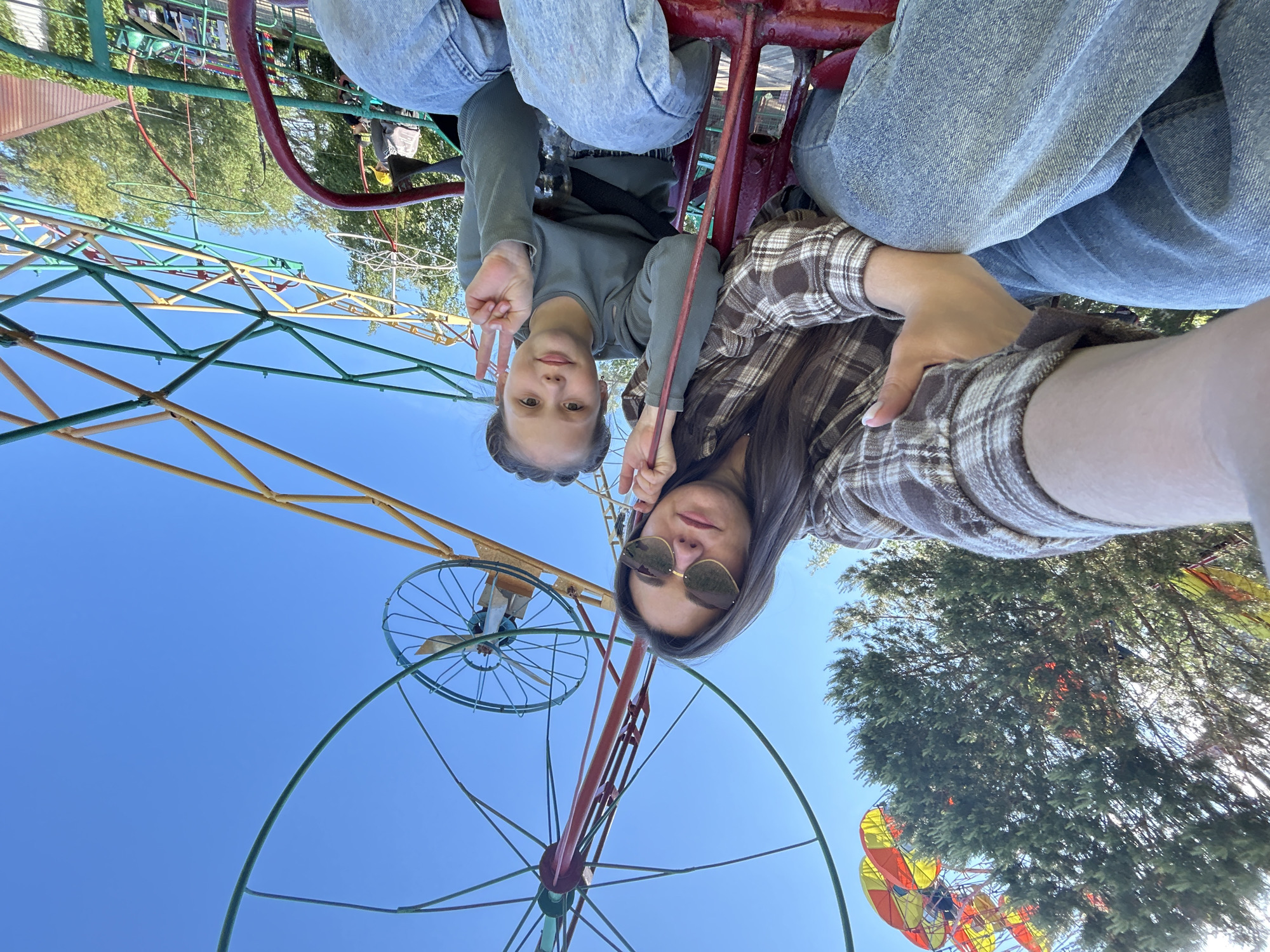 Two individuals are captured in a selfie while riding a Ferris wheel, with a view of the sky and partial park infrastructure in the background. The younger person gestures with two fingers. The older person wears sunglasses and a plaid shirt. They are seated on a structure that appears to be part of the Ferris wheel, and are presumably secured in the ride. The Ferris wheel structure itself is visible, with colorful steel beams and supporting framework. Behind the wheel, trees can be seen. The location is Chaikovsky, Russia.