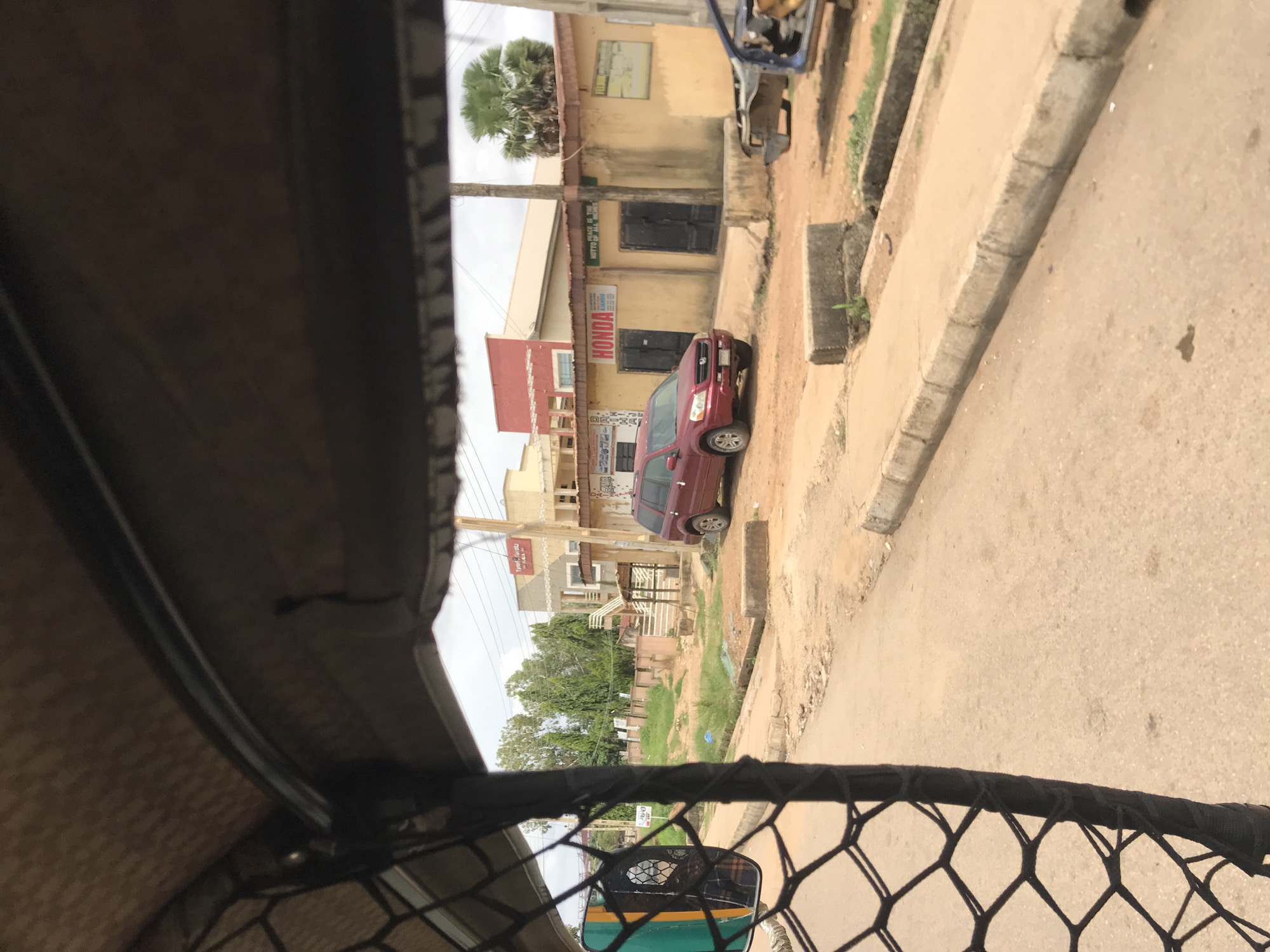 The image depicts a street scene in Rafin Par, Nigeria, viewed from the interior of a vehicle, likely a tricycle, as indicated by the dark brown fabric roof and a black, diamond-patterned mesh on the left. On the left side, a green-framed side mirror reflects an indistinct figure possibly wearing a plaid shirt.
The street is unpaved, consisting of packed dirt and gravel with visible cracks and uneven patches. Concrete curbs define the edges of the road, some of which are broken or show signs of wear. Small patches of green vegetation, including grass and weeds, grow alongside the curbs.
In the midground, on the right side of the street, a dark red first-generation Honda CR-V (circa 1997-2001) is parked, facing towards the left. Further down the road on the right, the front chassis and partial remains of a blue vehicle are visible, appearing to be stripped or derelict.
The background features several buildings under an overcast sky. On the right, a single-story building with a faded yellow-beige facade displays a prominent "HONDA" sign above two dark, closed doors. To its right, another sign reads "PEACE IS THE MOTTO OF ALL HOMES." A tall, dark utility pole stands directly in front of this building. Beyond this structure, a large palm tree with green fronds is visible. In the center-left background, a two-story building with a light-colored facade features a distinctive red section on its upper left portion and an external staircase leading to the second level. Another sign, partially obscured, is visible on this building. To the left and behind these structures, a cluster of tall, slender trees with dense green foliage forms a backdrop.