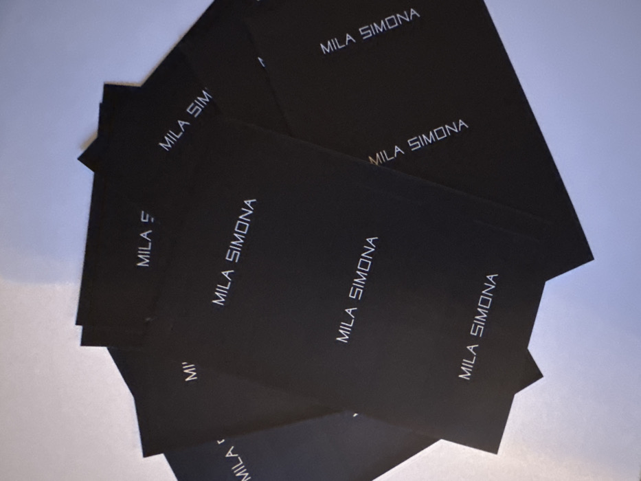 A collection of approximately ten to twelve black, rectangular cards or small brochures are fanned out and overlapping on a plain, light grey or white surface. Each visible card prominently displays the text "MILA SIMONA" in a white, sans-serif, uppercase font. The lettering exhibits a slightly futuristic or geometric style. The cards are arranged in a casual, somewhat scattered pile, with multiple instances of the full "MILA SIMONA" text visible at various angles, alongside partially obscured text on underlying cards. The lighting is diffuse and even, suggesting an indoor environment, casting subtle shadows from the edges of the cards onto the underlying surface. The scene is located indoors within the city of London, United Kingdom. No people, specific actions, or further environmental details are discernable beyond the immediate arrangement of the cards and the plain background.