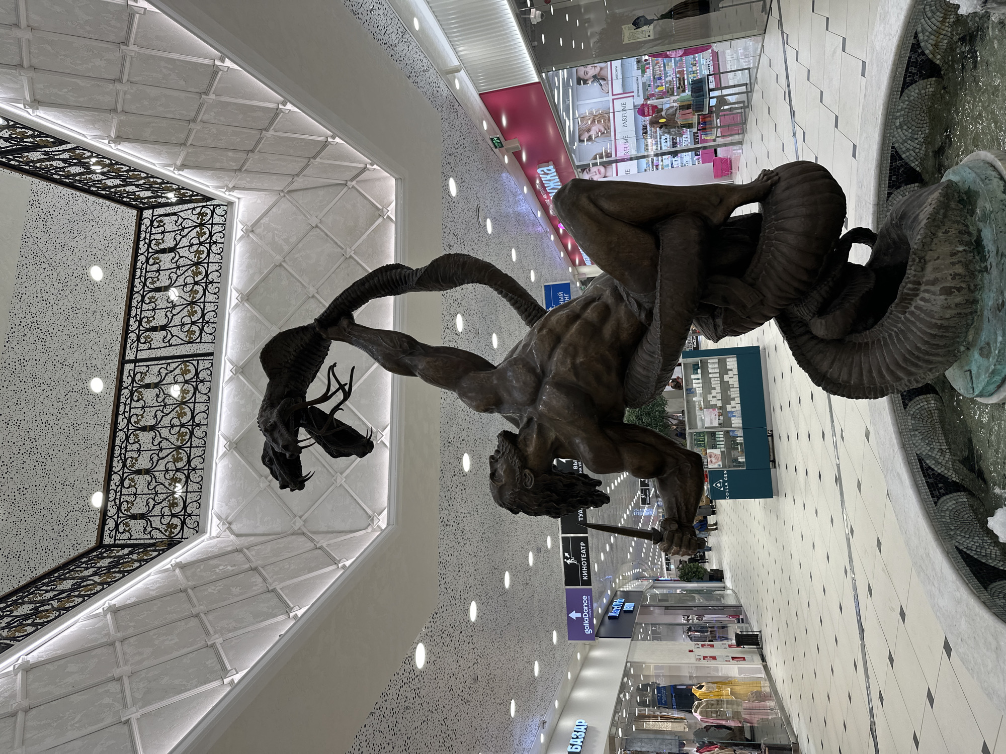 This image captures a grand bronze sculpture within a spacious, modern shopping mall in Yekaterinburg, Russia. The central subject is a large, muscular male figure, possibly a hero from mythology, engaged in a dramatic struggle with a multi-headed serpent or hydra. The statue is intricately detailed, with the man holding a spear or staff, poised to strike. The serpent's bodies are intertwined, some heads featuring prominent ram-like horns. This sculpture appears to be the centerpiece of a fountain, as water is visible in a decorative, mosaic-lined basin at its base.
The mall interior is well-lit and features a high ceiling with varied architectural designs, including coffered panels and speckled surfaces, enhanced by integrated LED strip lighting. The floor is a clean, tiled pattern of light and dark squares. Shops with glass storefronts line the walkways, displaying products like cosmetics and apparel. On the left, an ornate black and gold metal railing hints at an upper level or balcony.
Visible text on store signs includes "Рив Гош" (Rive Gauche), "ПАРФЮМЕРИЯ" (Perfumery), "gallaDance", "КИНОТЕАТР" (Cinema) with a directional arrow, and "БАЗАР" (Bazaar). Another sign reads "COLLA GEN". A few indistinct individuals can be seen in the background, walking through the mall. The overall scene suggests a vibrant commercial environment, likely bustling with shoppers, although the immediate area around the statue appears relatively clear. The indoor setting provides consistent bright lighting, making the time of day ambiguous.