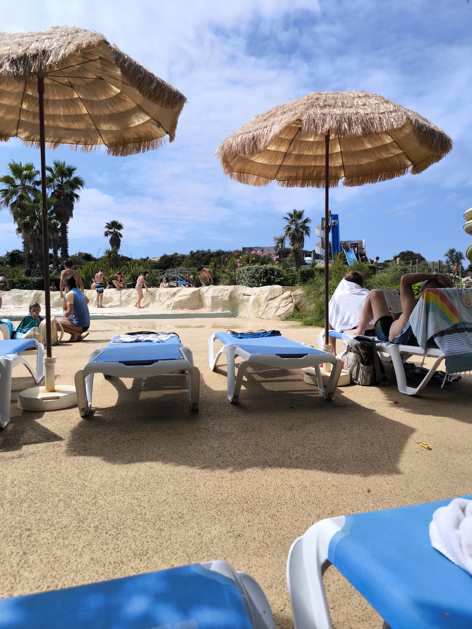The image depicts an outdoor scene at a water park, likely the Atlantic Park in Seignosse, France, on a sunny day. In the foreground, several blue and white lounge chairs are visible, some with towels draped over them. Two large, straw-thatched umbrellas provide shade.
In the mid-ground, there is a shallow water area with a rocky, beige-colored edge. Several people are present in this area, some wading or playing in the water, while others are sitting or standing nearby. Behind the water area, a blue water slide is prominent, with a spiral staircase leading up to it. More people are seen in the background, some appearing to be queuing for the slide or observing others. Palm trees and lush greenery dot the landscape, suggesting a warm climate. The sky is blue with scattered white clouds.
The overall impression is one of a recreational area designed for leisure and water activities. The focus is on the amenities and the presence of people enjoying themselves in a vacation-like setting.