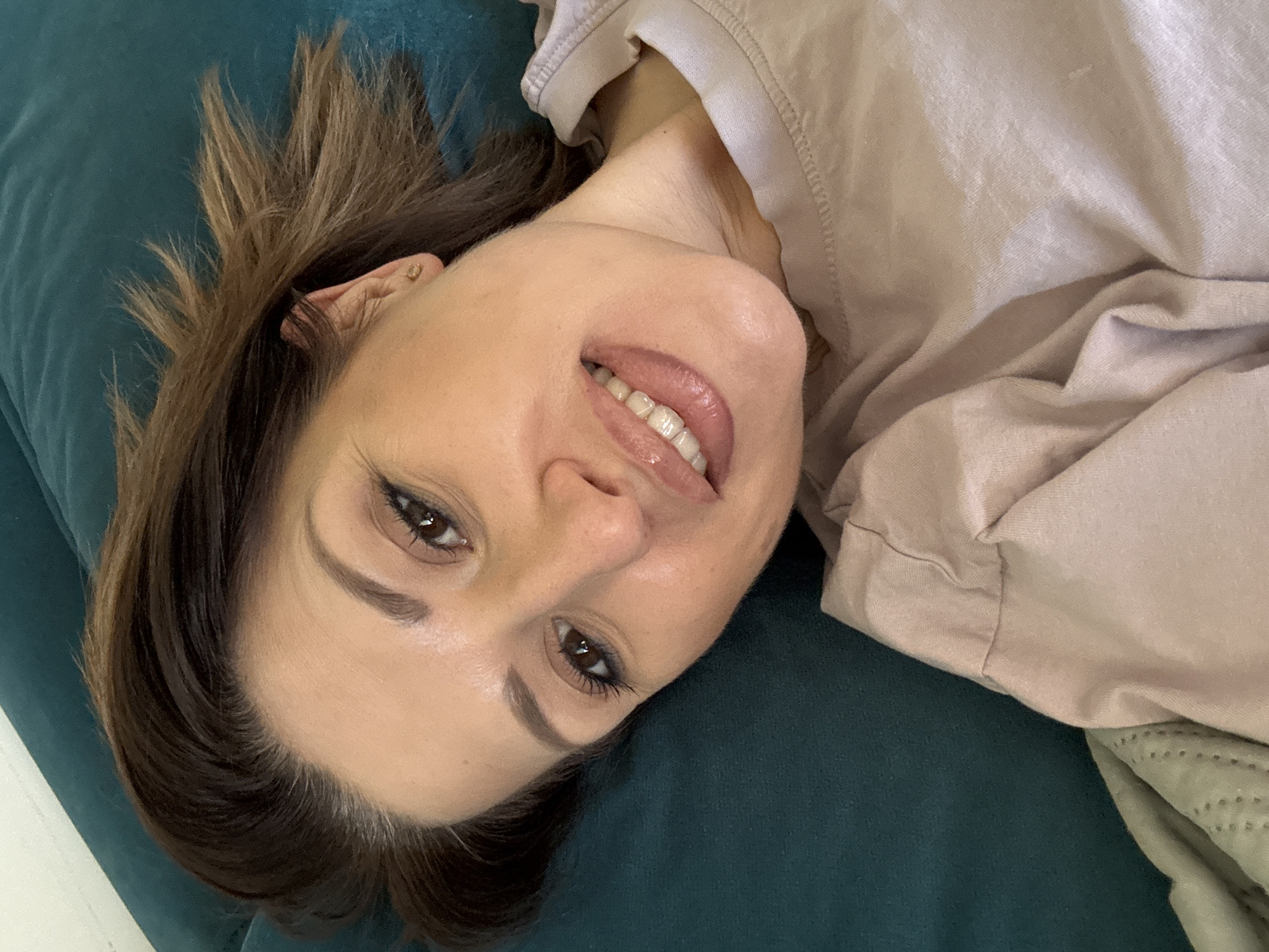 A woman is lying on a dark green velvet-like couch or cushion. She is fair-skinned, with light brown hair. She is wearing a light beige or pink long-sleeved shirt or sweater. She is smiling, with her teeth visible. She is indoors. The image doesn't give any information regarding the location in the city of Sofia, Bulgaria.