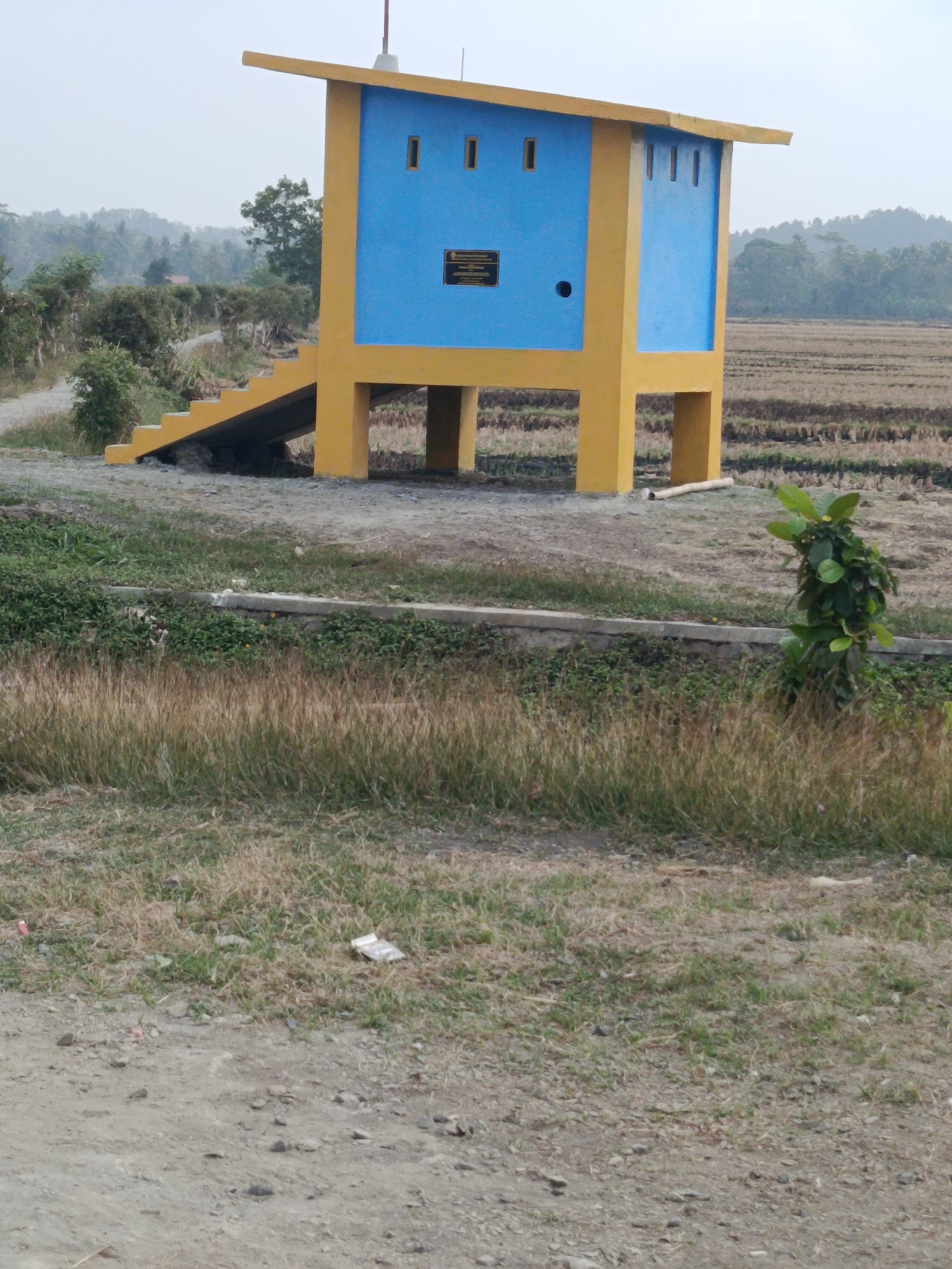 A yellow and blue elevated structure stands in a rural landscape. The structure has a yellow base and legs supporting a blue cuboid with a yellow roof. The blue section has small rectangular openings and a circular opening on its side. A yellow staircase leads up to the structure. In the background, a dry agricultural field is visible, with trees and a hazy, possibly mountainous, landscape beyond. The foreground consists of a dirt path and dry grass. The overall impression is one of a remote or developing area. The location is identified as Bantarsari, Indonesia.