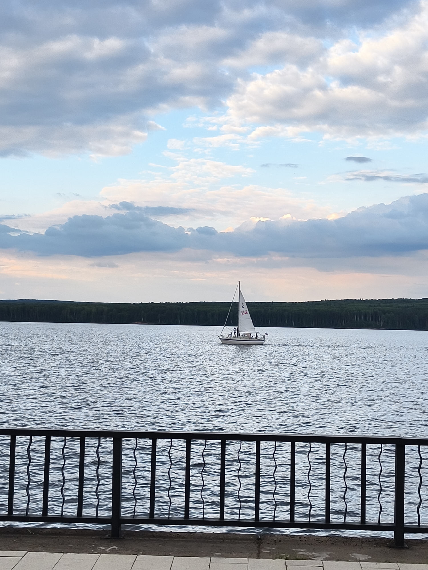 A sailboat is gliding across a body of water, possibly a lake or a large river, under a cloudy sky. The sky is a mix of pale blue and various shades of gray clouds, some appearing fluffy and white, others darker and more ominous. The water's surface is rippled, reflecting the overcast sky.
In the distance, a dense line of dark green trees forms the horizon, suggesting a forested area bordering the water. In the foreground, a black metal fence with decorative wavy vertical bars separates the viewer from the water. The fence is set against a paved walkway and a patch of ground.
On the sailboat, several people are visible, suggesting a leisurely outing or a sailing activity. The boat has a white sail with some markings on it. The overall atmosphere is calm and serene, with the time of day appearing to be late afternoon or early evening, given the soft light and the mix of light and dark clouds. The scene evokes a sense of peace and outdoor recreation. There is no visible text in the image.