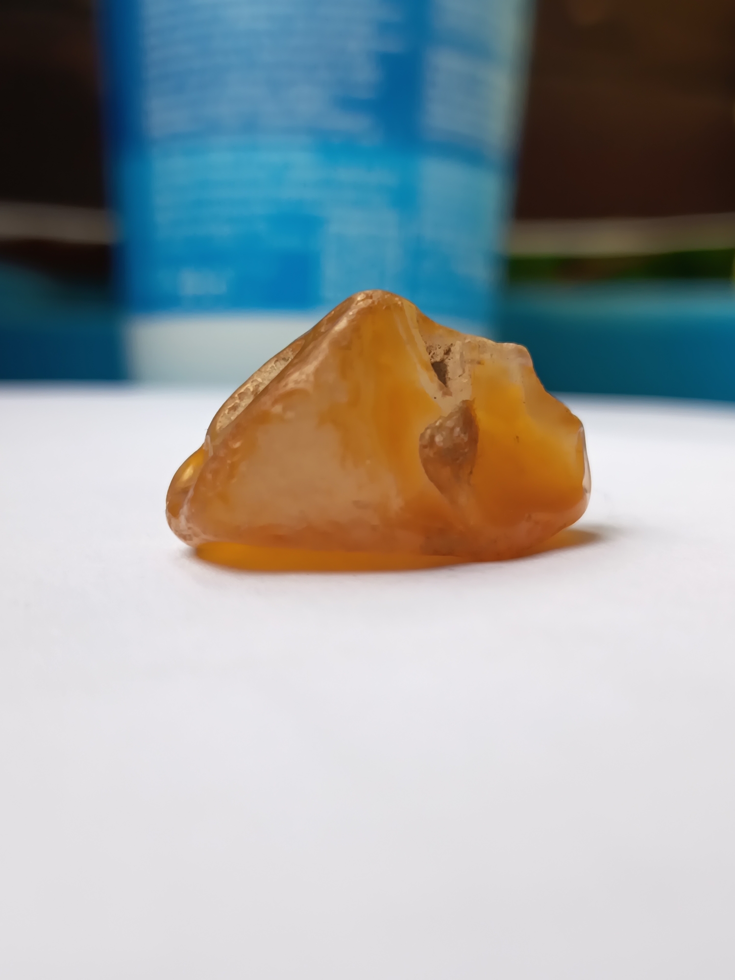 A close-up, low-angle, eye-level shot focuses on a single piece of amber, which is translucent and orange-yellow in color. The amber appears rough and irregularly shaped, with a cavity visible in its surface. It rests on a white, flat surface. In the background, out of focus, are elements that suggest an indoor environment with some brightly colored objects, possibly plastic containers, one of which has a blue hue. There are no people or animals present in the image. The setting does not provide any specific geographical markers that would indicate a location within the city of Izmail, Ukraine.