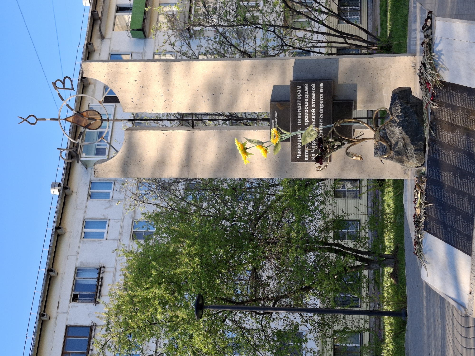 This is an outdoor shot of a memorial in the city of Chaikovsky, Russia, likely honoring those who died in the Chernobyl disaster. The monument is made of concrete and features a metal sculpture at the top consisting of a circle with elements resembling the letters "В" and "Ю". A smaller plaque reads in Russian, "To the people of Chaikovsky, liquidators of the catastrophe that occurred at the Chernobyl atomic station on April 26, 1986." Yellow flowers are placed beneath the plaque. To the right of the plaque, there is a large stone. Another plaque with names listed is also present. The monument is set against a building with multiple windows, and a tree is in the foreground. The sky is blue.