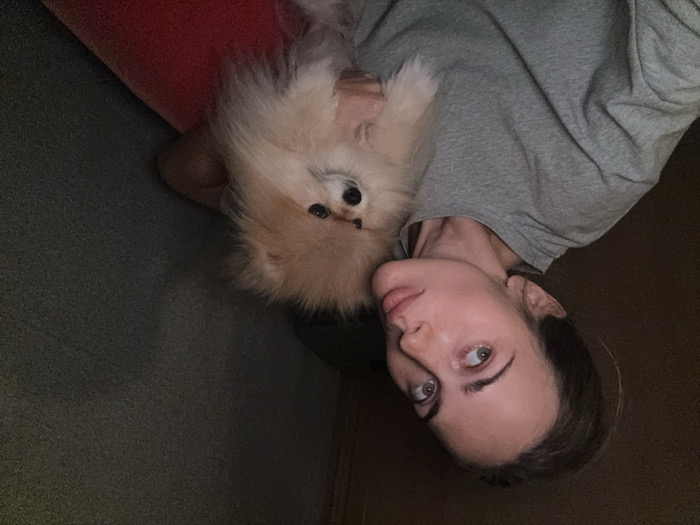The scene depicts an adult female and a small dog indoors, in Warsaw, Poland. The female, positioned on the left, has fair skin, light-colored eyes, and dark hair pulled back. She wears a grey long-sleeved top and her lips are pursed. She holds a fluffy, light brown Pomeranian dog with dark eyes in her arms. Both subjects are looking generally towards the camera. The environment is dimly lit, featuring a dark brown wall behind the female and a dark grey, textured surface to her right. A red object, possibly a cushion or blanket, is partially visible in the lower right corner.