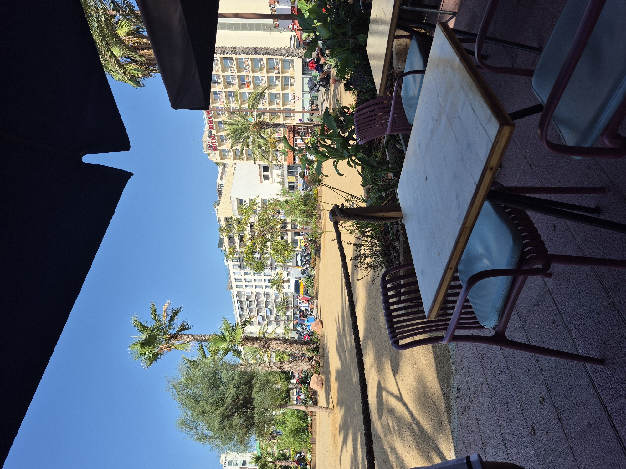 This is a photograph taken outdoors, possibly from a cafe or restaurant, looking towards a street and a beach area in Lloret de Mar, Spain. In the foreground, there are two tables with chairs. The tables appear to be made of wood and have metal legs. The chairs are a light purple color with light blue cushions.
Beyond the cafe area, there is a sandy area with some small rocks and a rope barrier. Several palm trees and other greenery are visible. In the background, there is a street with cars, motorcycles, and pedestrians. A large building with "HOTEL ROSAMAR" in red letters is prominent. The sky is clear and blue, indicating a sunny day.