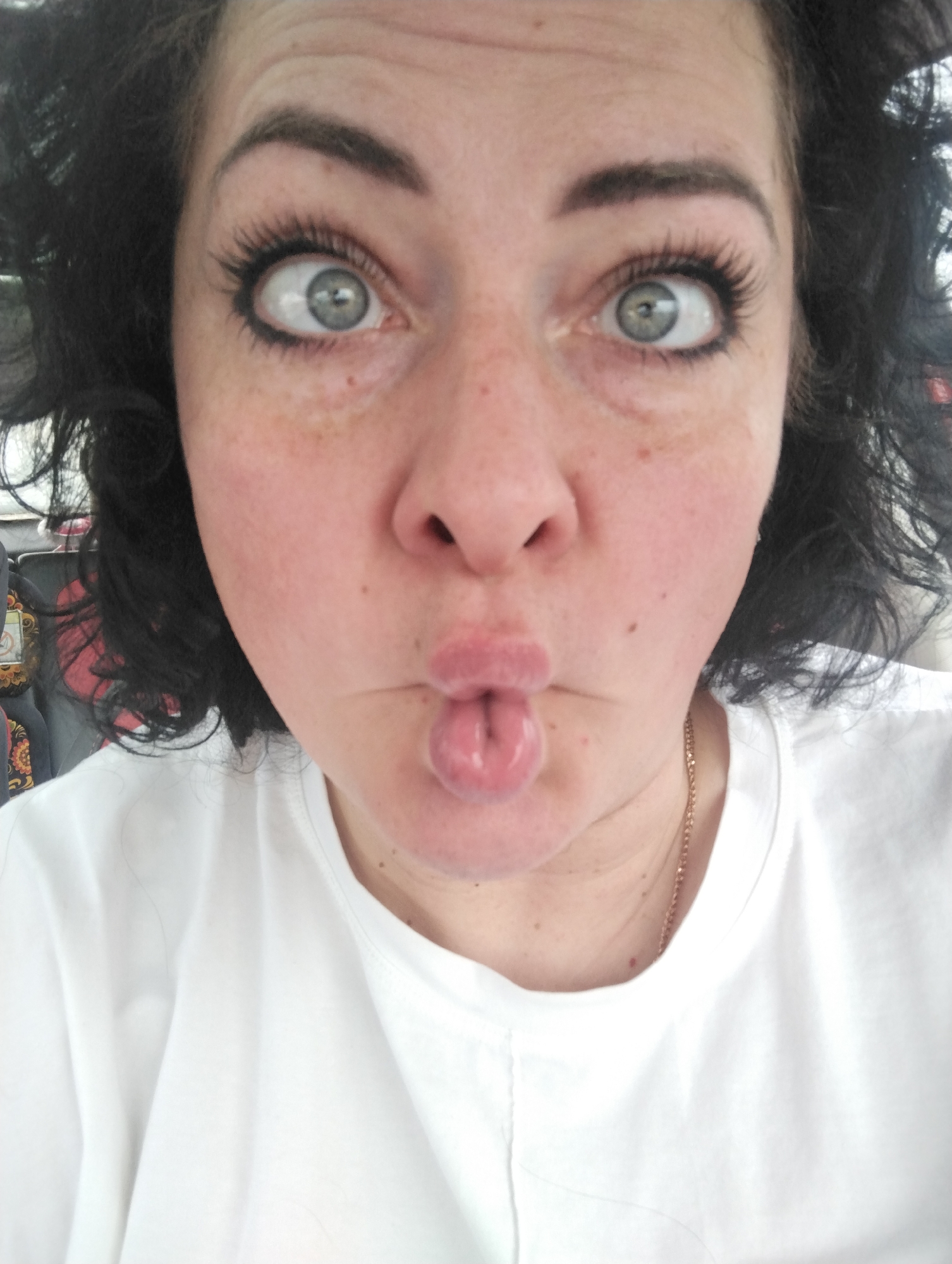 A close-up, front-facing portrait captures a person from Izhevsk, Russia, making a playful facial expression. The subject has medium-length, dark, curly hair visible on either side of their face. Their eyes, wide open, are a striking light blue or grey-green, with prominent dark eyelashes and well-defined eyebrows. There are freckles visible across their nose and cheeks. The mouth is pursed into a "fish lips" pose, with the tip of the tongue slightly visible between the lips, contributing to a humorous and lighthearted mood. The person is wearing a simple white t-shirt, and a thin, gold-colored chain necklace is faintly visible around their neck. The lighting suggests an indoor setting. No specific text is visible in the image.