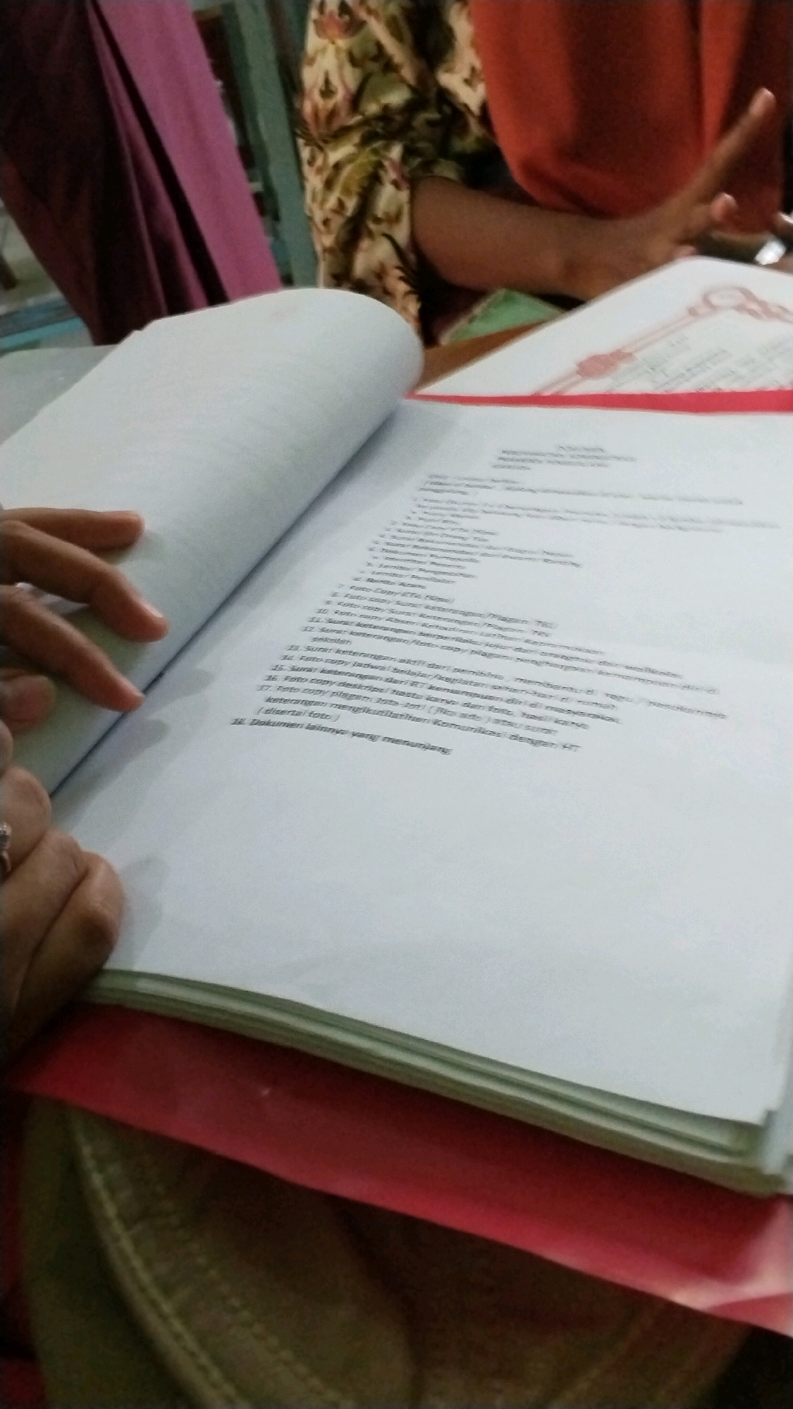 The image shows a close-up of a document being held open by a person's hand, with a red folder beneath it. The document appears to be a list of requirements or a checklist, with numbered items. Some of the text is legible, including the phrases "Surat keterangan dari..." and "Foto copy piagam, foto kopi...". The background shows parts of people's clothing and what appears to be a table. The lighting suggests an indoor setting. The context provided about Banjaragung, Indonesia, is external information and cannot be inferred from the image itself.