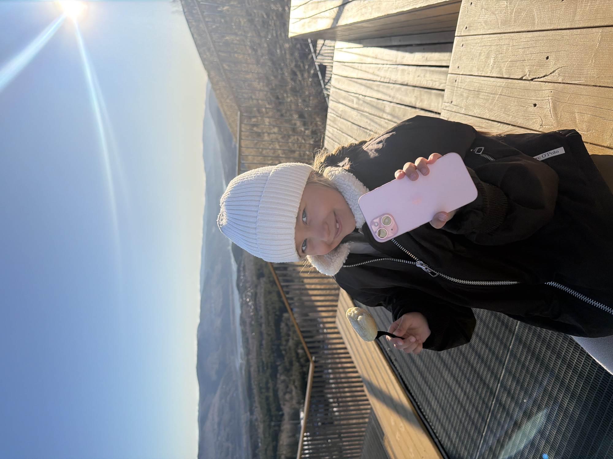 A young girl wearing a white beanie and a black jacket stands on an observation deck in the Stolby Nature Reserve near Krasnoyarsk, Russia. She holds a pink cell phone in one hand and a marshmallow on a stick in the other. In the background, a mountainous landscape stretches towards the horizon under a clear, bright sky. The observation deck is made of wood, and sections are made of metal grating.