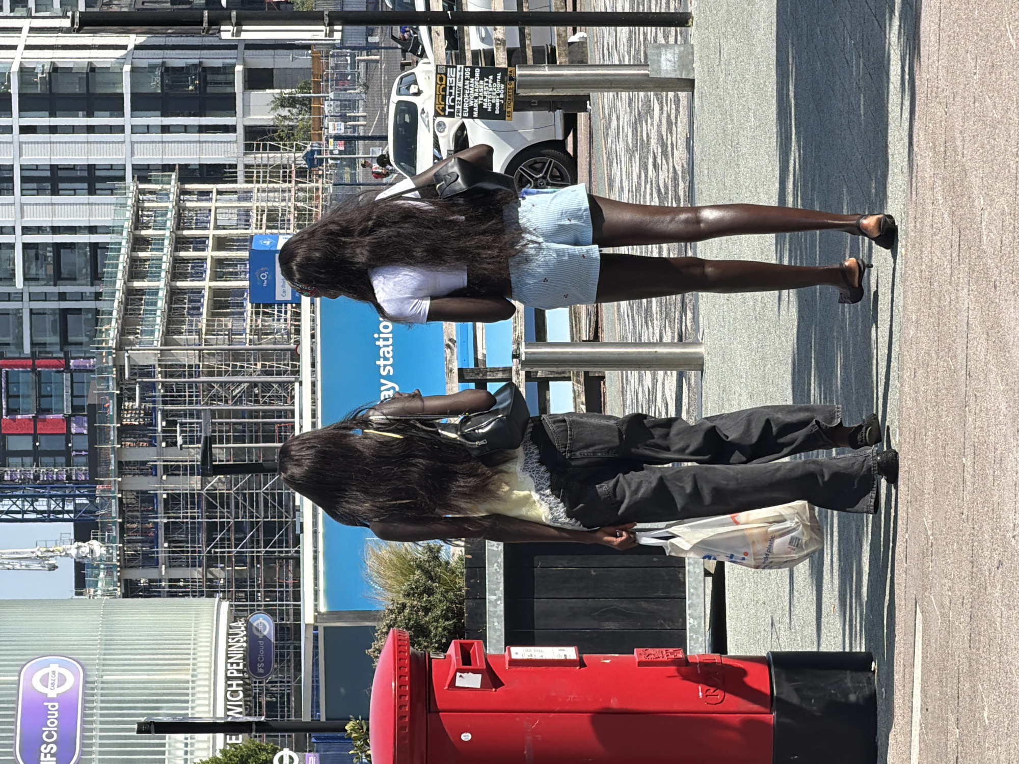 The image captures two Black women standing in front of a red British post box in the city of London, United Kingdom. One woman wears a white t-shirt and blue striped shorts, and high heels and carries a black purse. The other woman wears a yellow top and black jeans and holds a white plastic bag. In the background, there's a building under construction, a white car, and a sign for "IFS Cloud Cable Car - Greenwich Peninsula." A sign for "Afro Tribe" is visible. The location appears to be a station or transportation hub.