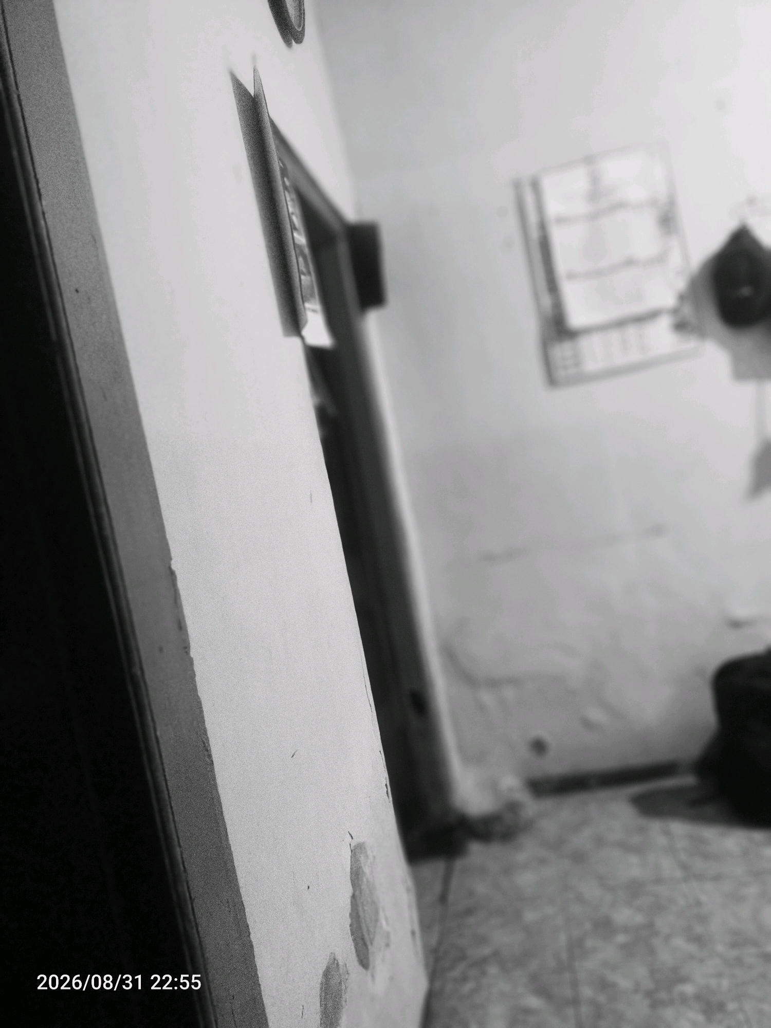 The image shows an interior scene, captured in black and white. In the foreground, on the left, is a textured surface that appears to be a wall or doorframe, angled towards the viewer. A portion of a doorway is visible to the right of this foreground element. The background is out of focus but suggests a room with a light-colored wall and a patterned floor. On the wall in the background, there is a framed item, possibly a calendar or a picture, and further to the right, what appears to be a dark-colored bag. The bottom left corner of the image displays a timestamp: "2026/08/31 22:55". The location of Jati Wates, Indonesia, is not discernible from the visual information provided in the image.