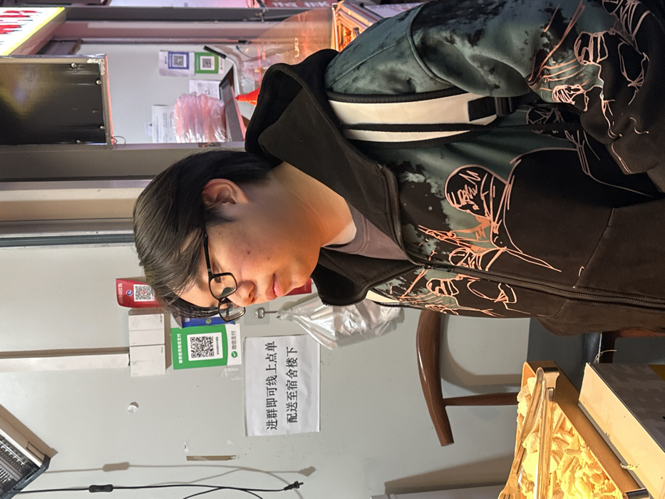 An East Asian male stands within a food establishment in Jiangjin, China. The individual, positioned slightly left of center, possesses dark, medium-length hair and wears black-framed eyeglasses. He is dressed in a black hooded zipper jacket, adorned with a teal/green and black patterned design featuring orange-red outlined figures, and a grey inner collar. A white backpack strap is visible over his left shoulder. He maintains a neutral expression, gazing towards the right.
The background on the left features a light-colored wall with a wall-mounted heater in the upper left. Below this, electrical wiring and a white paper sign with black Chinese characters are present. The sign reads "进群即可线上点单 配送至宿舍楼下" (Join the group to order online, delivered downstairs to the dormitory). Above the sign are a green QR code for "微信支付" (WeChat Pay) and a red QR code. A wooden chair with a curved backrest is partially visible behind the subject. In the foreground, a metal display tray contains various fried snack items, accompanied by metal tongs. The right background, partially out of focus, reveals additional display shelving with wrapped items and another QR code, indicative of a commercial setting.