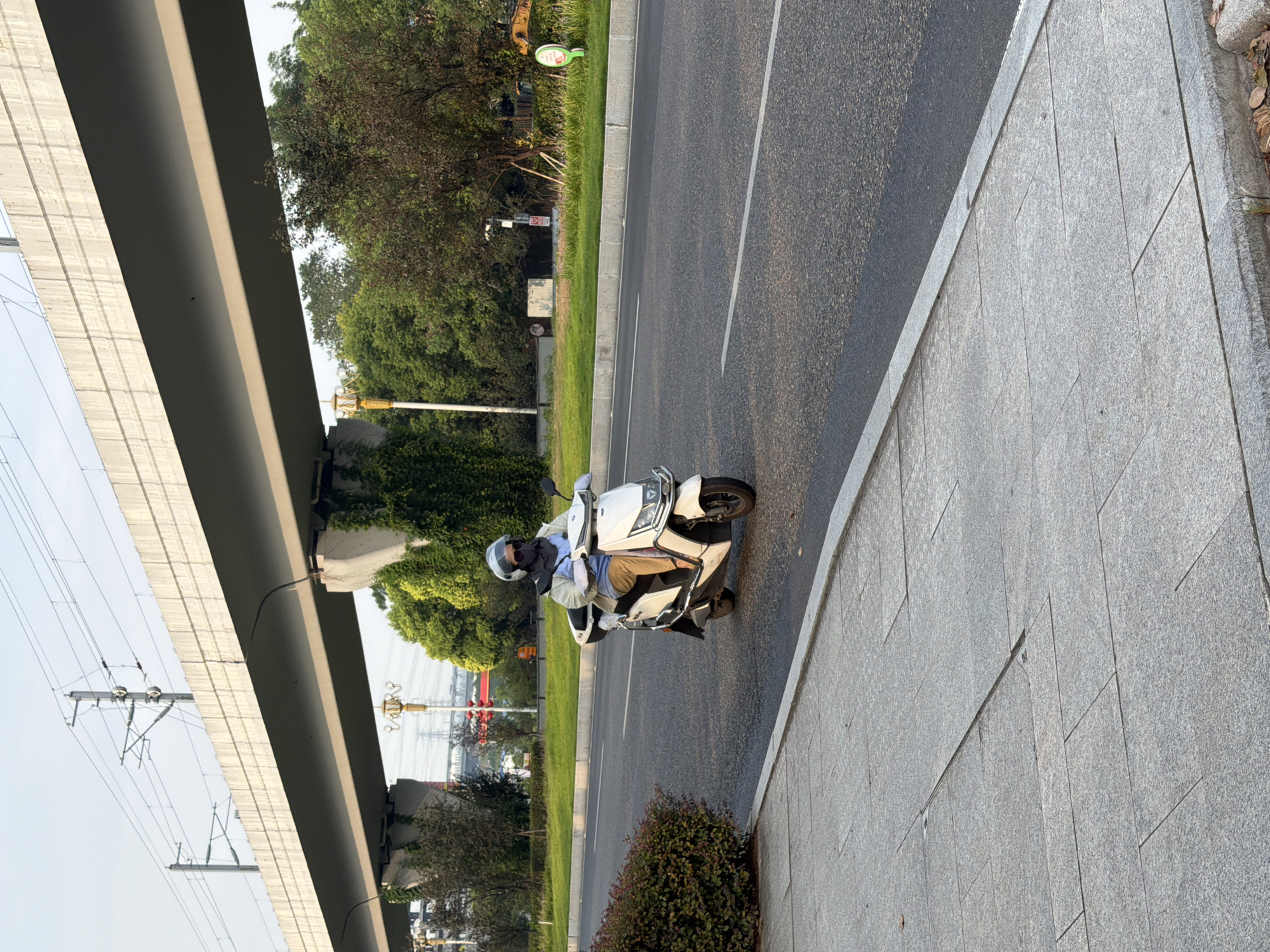 A person wearing a helmet, a face mask, and a blue shirt with tan pants rides a white electric scooter along a road in Jiangjin, China. The scooter is in motion, facing towards the right of the frame. In the background, a large elevated structure with overhead power lines spans across the scene. Trees and greenery line the side of the road, and a sidewalk is visible in the foreground. The sky is overcast.