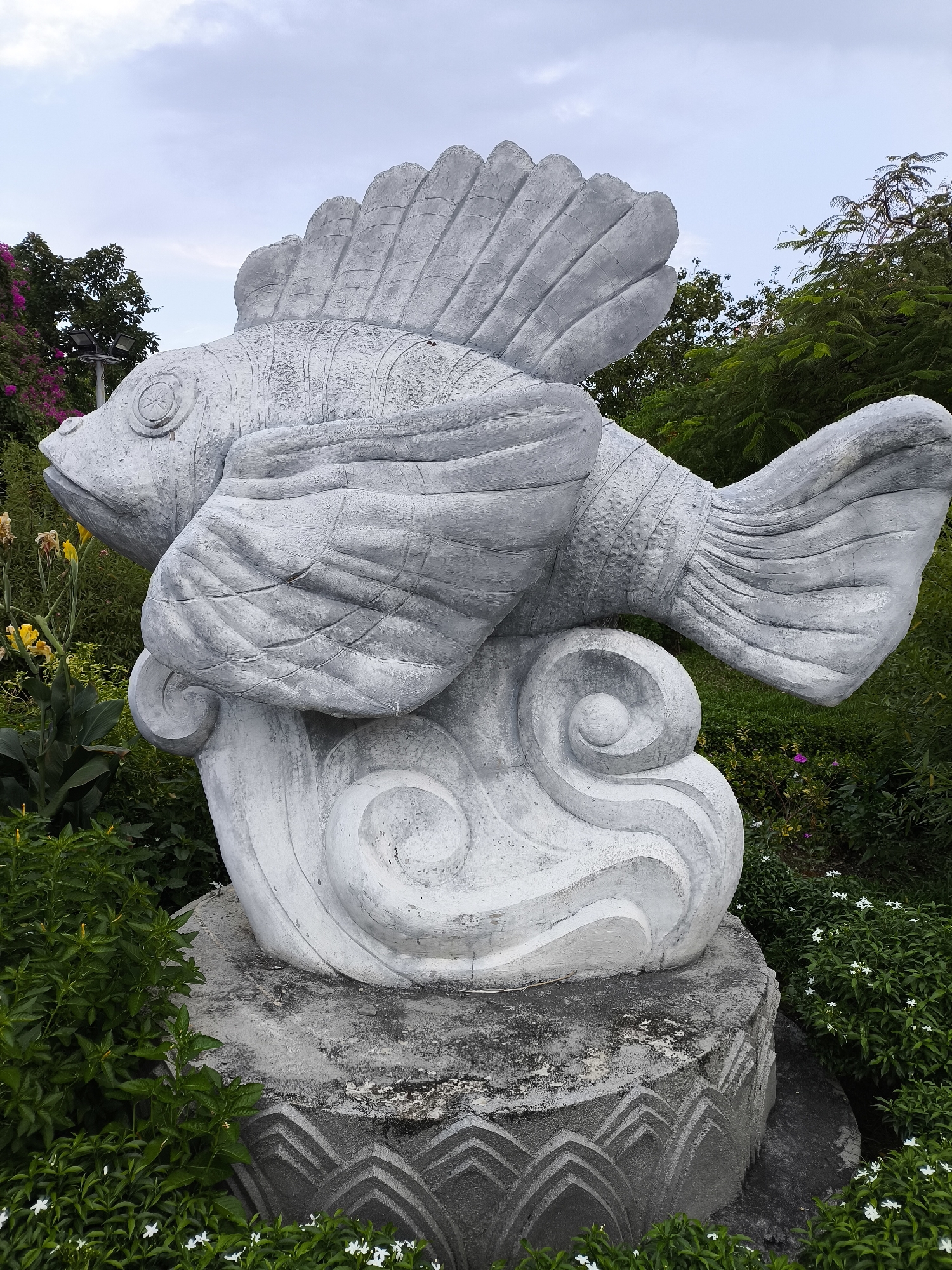 A grey, weathered concrete sculpture of a stylized fish is the central subject of the image. The fish is positioned mid-leap, with its body curving upwards and its fins spread. It rests on a base that resembles stylized waves, also made of concrete. The sculpture sits in an outdoor setting, surrounded by lush green foliage and some flowering plants. In the background, trees and a cloudy sky are visible. The base of the sculpture has a decorative, stepped pattern. The overall impression is of a monument or decorative element in a garden or public space. The location is identified as Bac Nha Trang Ward, Vietnam. There are no people visible in the image.