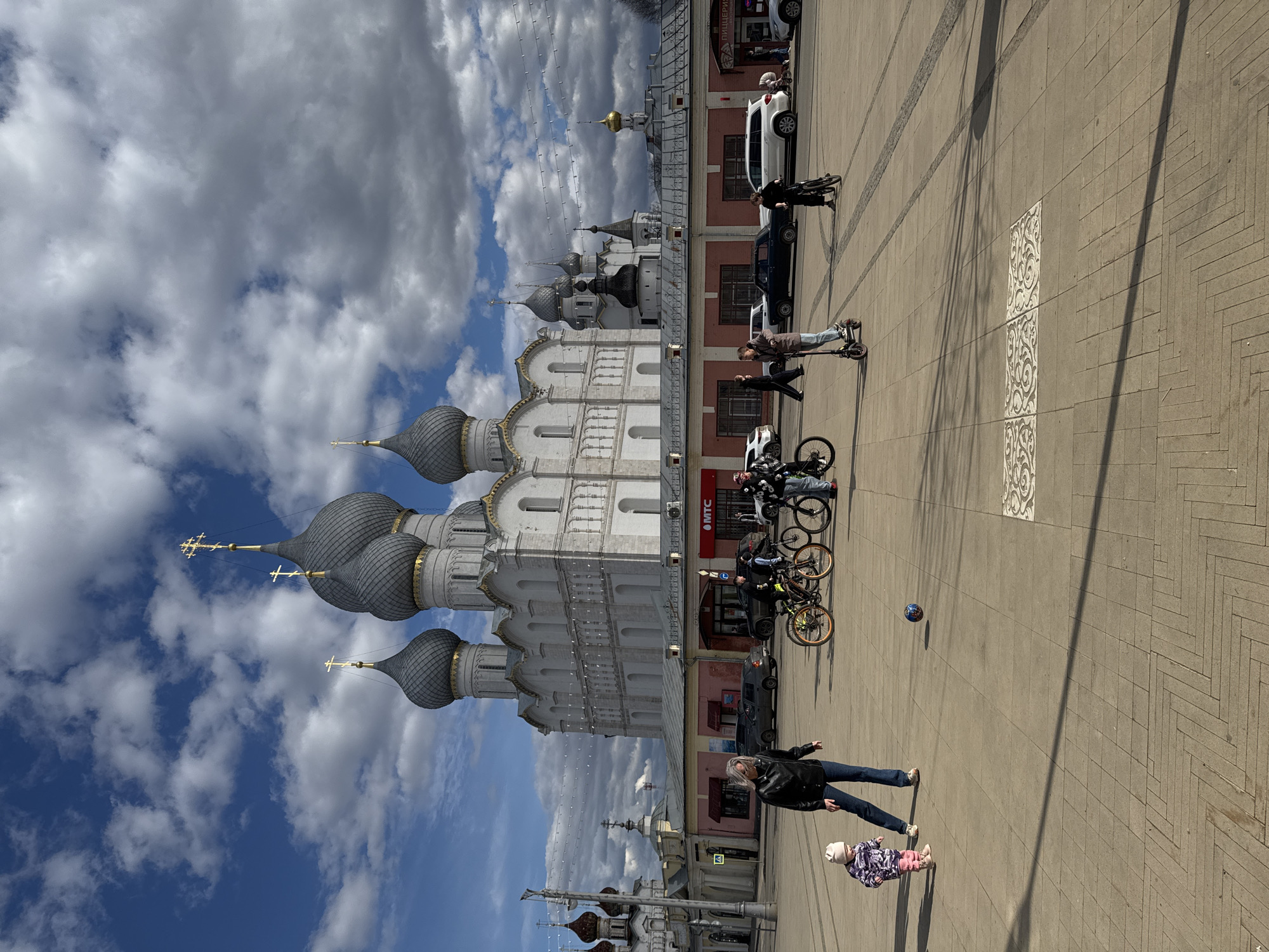 The image shows a street scene in Семибратово, Russia, dominated by a white building with multiple grey onion domes topped with gold crosses under a sky with fluffy clouds. Adjacent to the building is a line of parked cars and commercial storefronts. Several people are visible: some are on bicycles, one is riding a scooter, and a woman is standing near a young child. A soccer ball is also visible on the pavement. The ground is paved with reddish-brown bricks. A shadow from a lamp post stretches across the pavement.