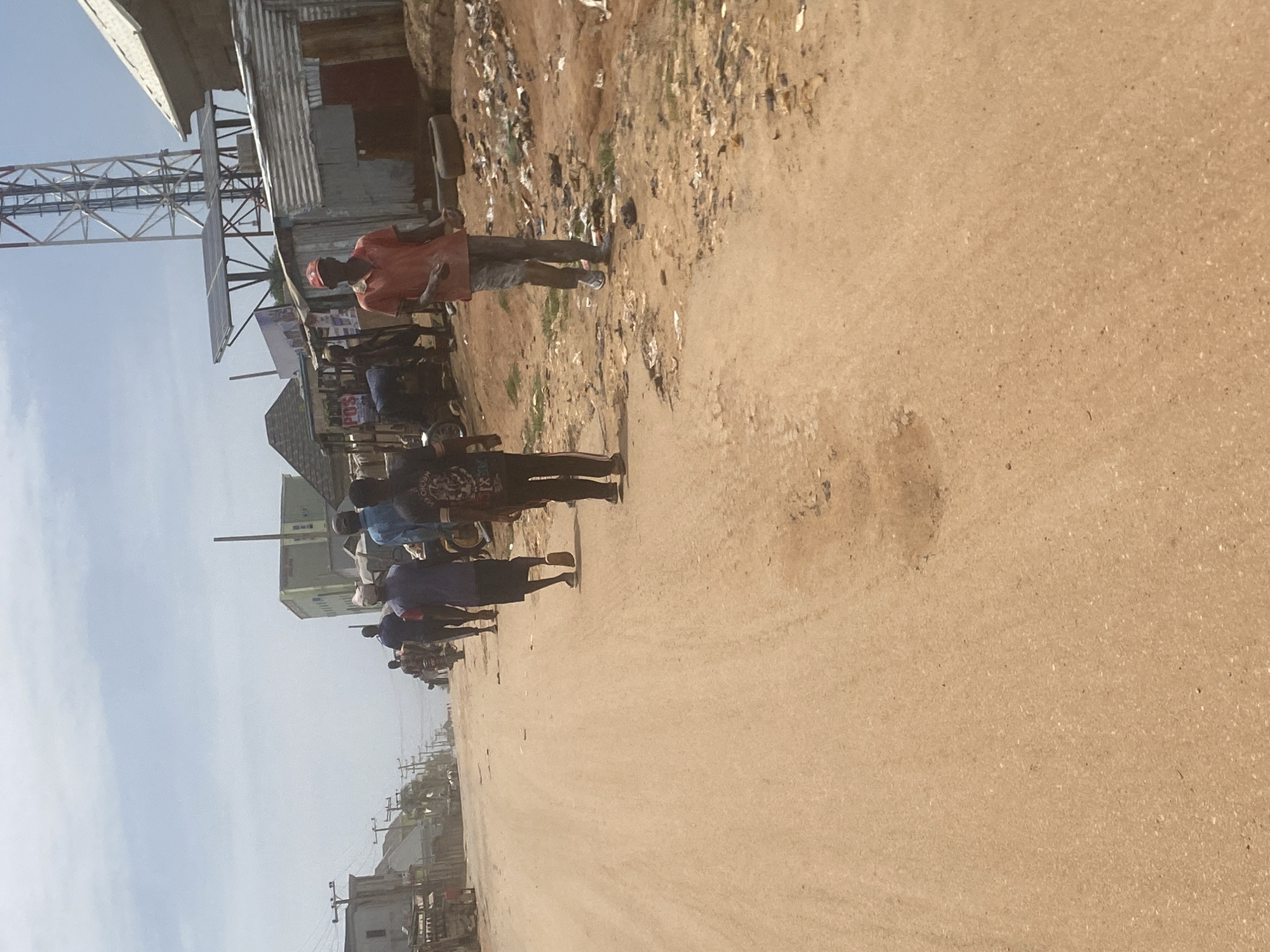 The image shows a street scene in Lugbe, Nigeria. A group of people are walking along the dirt road. There is trash along the side of the road. Buildings and a communication tower are visible in the background. The sky is overcast.