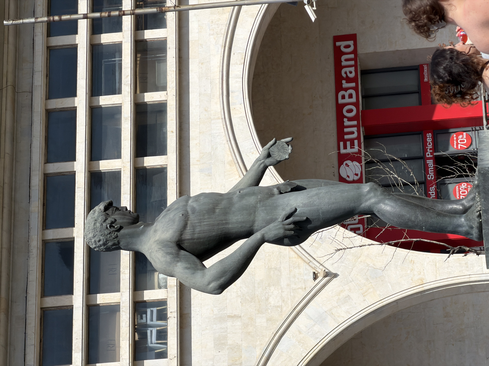 The image shows a bronze statue of a nude male figure holding a disc, standing in front of a building with a "EuroBrand" store. The building is located in Tbilisi, Georgia, and has a facade with arched openings and rectangular windows. Two people are seen on the right side of the frame, possibly interacting with the store. The "EuroBrand" store has red signage indicating "Small Prices" and a "70% off" discount.