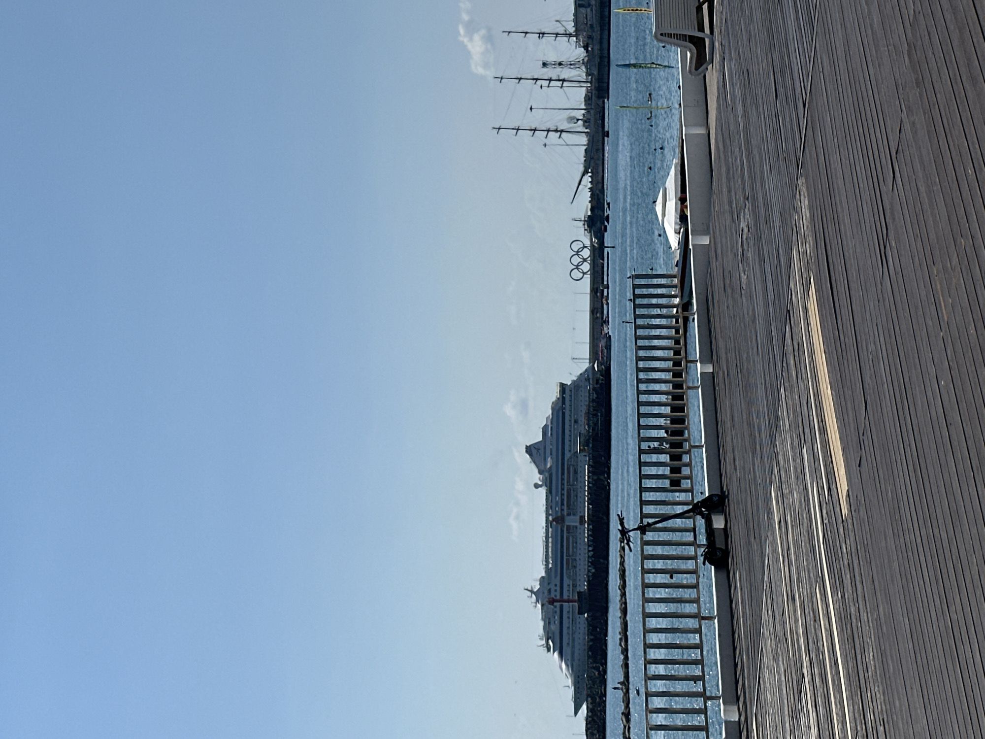 This image shows a clear, sunny day at the Sochi Sea Port in Sochi, Russia. In the foreground, a weathered wooden pier stretches across the bottom of the frame, with a dark grey scooter parked on it. A white metal railing runs along the edge of the pier, separating it from the blue water beyond. In the mid-ground, a large white cruise ship is docked, appearing to be the "AIDAblu." To the right of the cruise ship, the iconic Olympic rings are visible, situated above the water and near other docked ships, including several tall sailing vessels. The water is calm and a few people can be seen in and around the water in the distance. The sky is a clear, bright blue with a few wispy clouds.