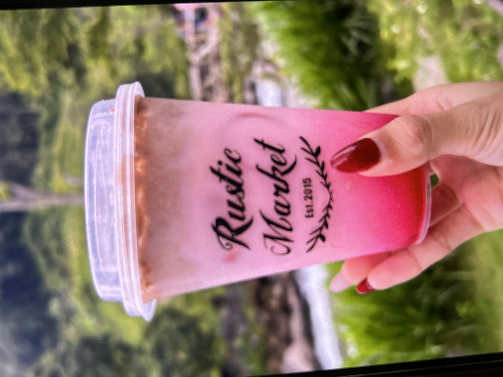 A hand with dark red nail polish holds a clear plastic cup filled with a pink layered drink, topped with a clear plastic lid. The cup has black text that reads "Rustic Market Est. 2015" with decorative branches on either side of the year. The background is blurred, showing lush green foliage and a body of water, suggesting an outdoor natural setting, possibly near a park or river in Depok, Indonesia. The lighting is soft and natural, consistent with daylight.
