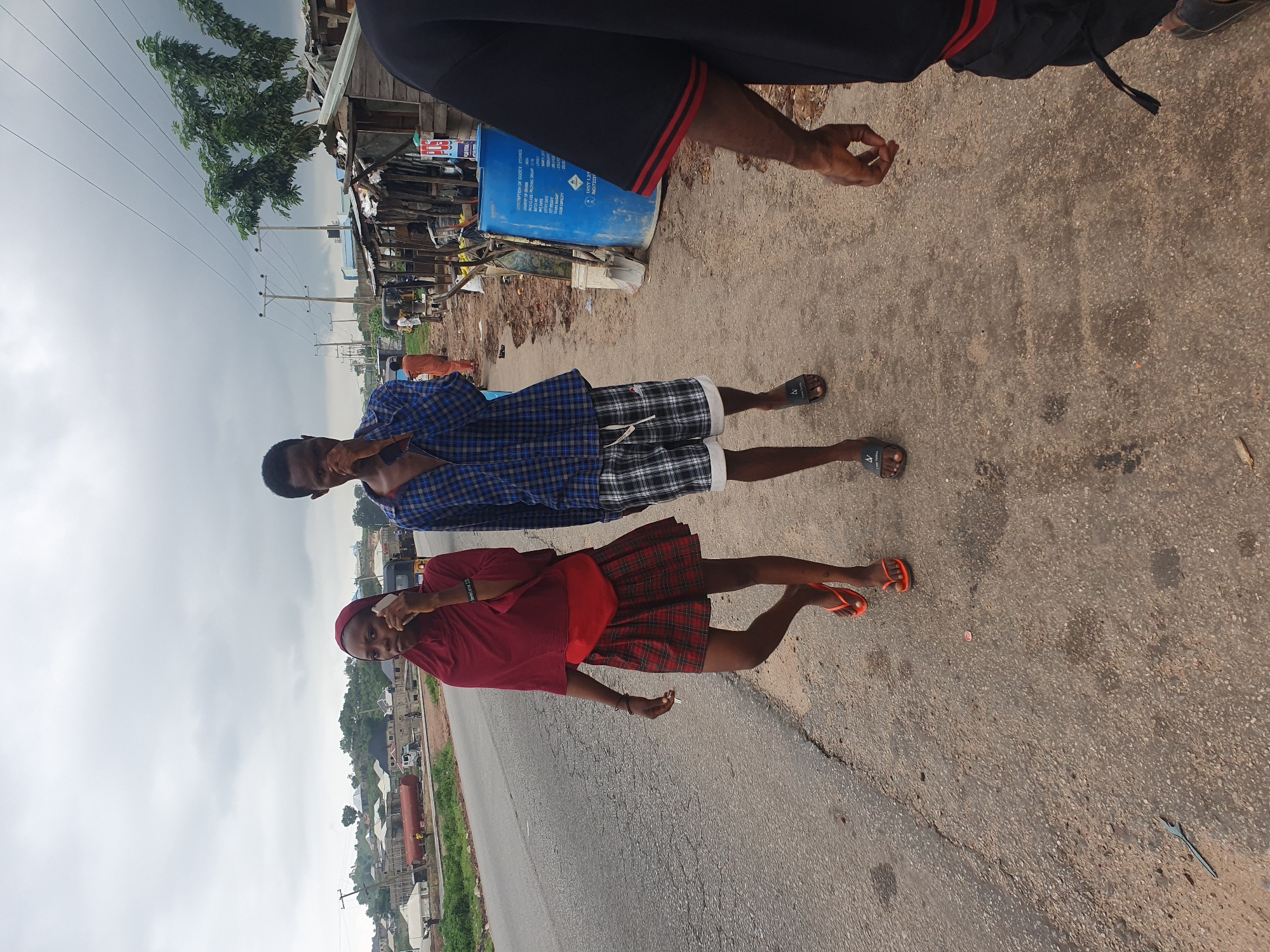 Two young people, a female wearing a maroon hijab and a red and black plaid skirt, and a male wearing a blue and black checkered shirt and plaid shorts, are walking on the side of a paved road. The female is holding a cell phone to her ear. In the background, there are makeshift market stalls with corrugated metal roofs, a blue plastic barrel, and trees. A truck and several houses are visible on a hill in the distance. The sky is overcast. This scene is consistent with the environs of Lokoja, Nigeria, a city known for its roadside markets and proximity to natural topography.
