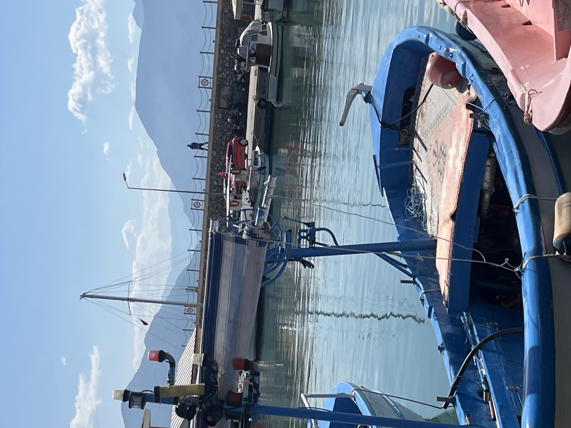 The image depicts a view in the city of Alanya, Turkey. In the foreground, the details of two docked fishing boats are visible, one painted blue and another pink. The blue boat contains nets and rope. The boats sit on calm, reflective water. A stone pier runs along the water, lined with a decorative railing featuring circular designs. A single person walks along the pier. Further back on the pier, various vehicles, including a red car and a white van, are parked. The background features a clear blue sky and distant, hazy mountains.