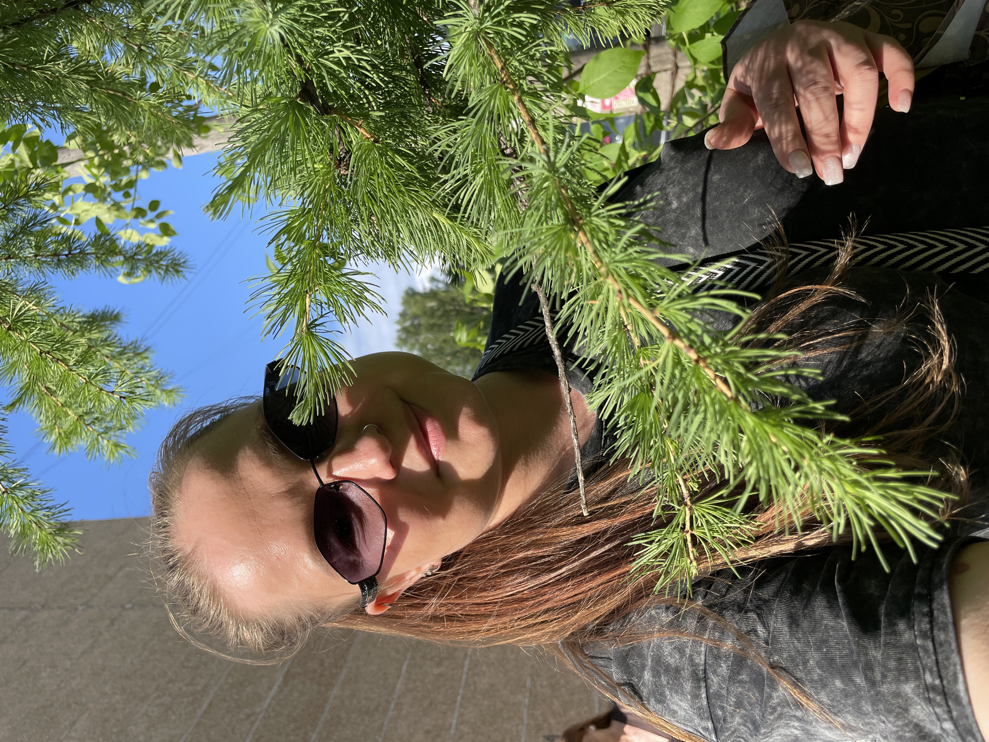 A woman with auburn hair and sunglasses poses for a photo beneath pine tree branches in what appears to be outdoor setting. The blue sky is visible behind the branches. Her hand is visible on the right, her nails manicured with white polish. The woman is wearing a dark jacket. The location, Izhevsk, Russia, cannot be confirmed from the image.
