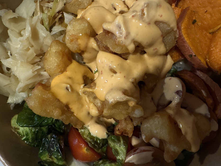 The image shows a close-up of a food bowl with various ingredients. There's fried food, possibly potatoes or tofu, generously drizzled with a creamy, reddish-orange sauce. To the side, there's a portion of sliced sweet potato. The bowl also contains shredded cabbage, Brussel sprouts, and halved cherry tomatoes. The dish appears to be a combination of sweet, savory, and fried components.