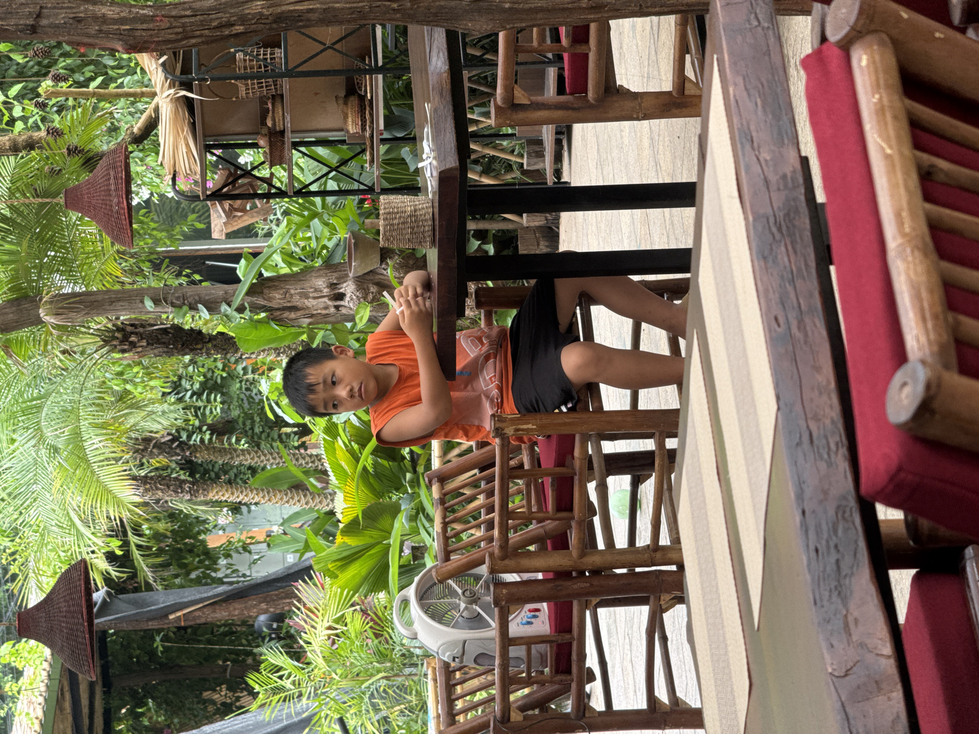 A young boy, approximately 8-10 years old, sits at a wooden table outdoors, facing to the right. He wears an orange sleeveless shirt and black shorts. His hands are clasped on the table. The table is set with two placemats and a small woven basket. Behind the boy, bamboo chairs with red cushions are visible, as is a white electric fan. The environment appears to be a restaurant or cafe with abundant tropical foliage, including palm trees. In the background, a shelf unit displays various woven items. The image is taken in Nam Hoa Lư Ward, Vietnam.