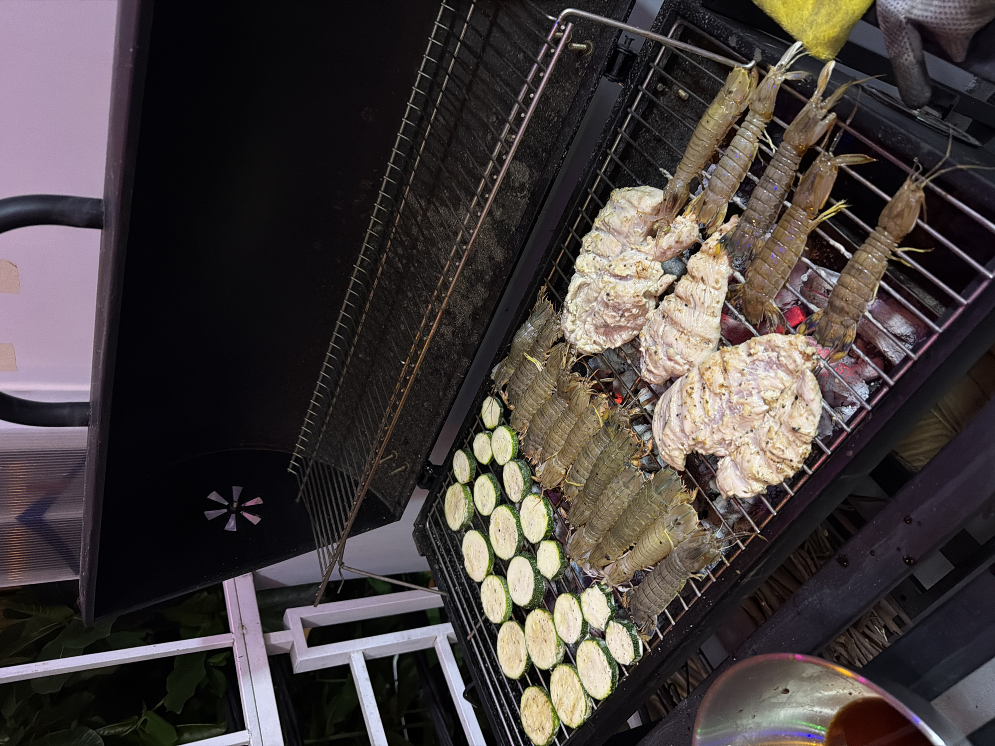Captured in Nha Trang Ward, Vietnam, the image presents a grill loaded with assorted food items. The grill is filled with sliced zucchini, mantis shrimps, and marinated meat. The zucchini slices appear to be seasoned and evenly distributed on the grill. Several mantis shrimps are laid out alongside the zucchini. The marinated meat is shaped into large, irregular portions. The grill itself is positioned in an outdoor setting, with a hint of foliage visible beneath it. The overall lighting suggests it is evening or night.