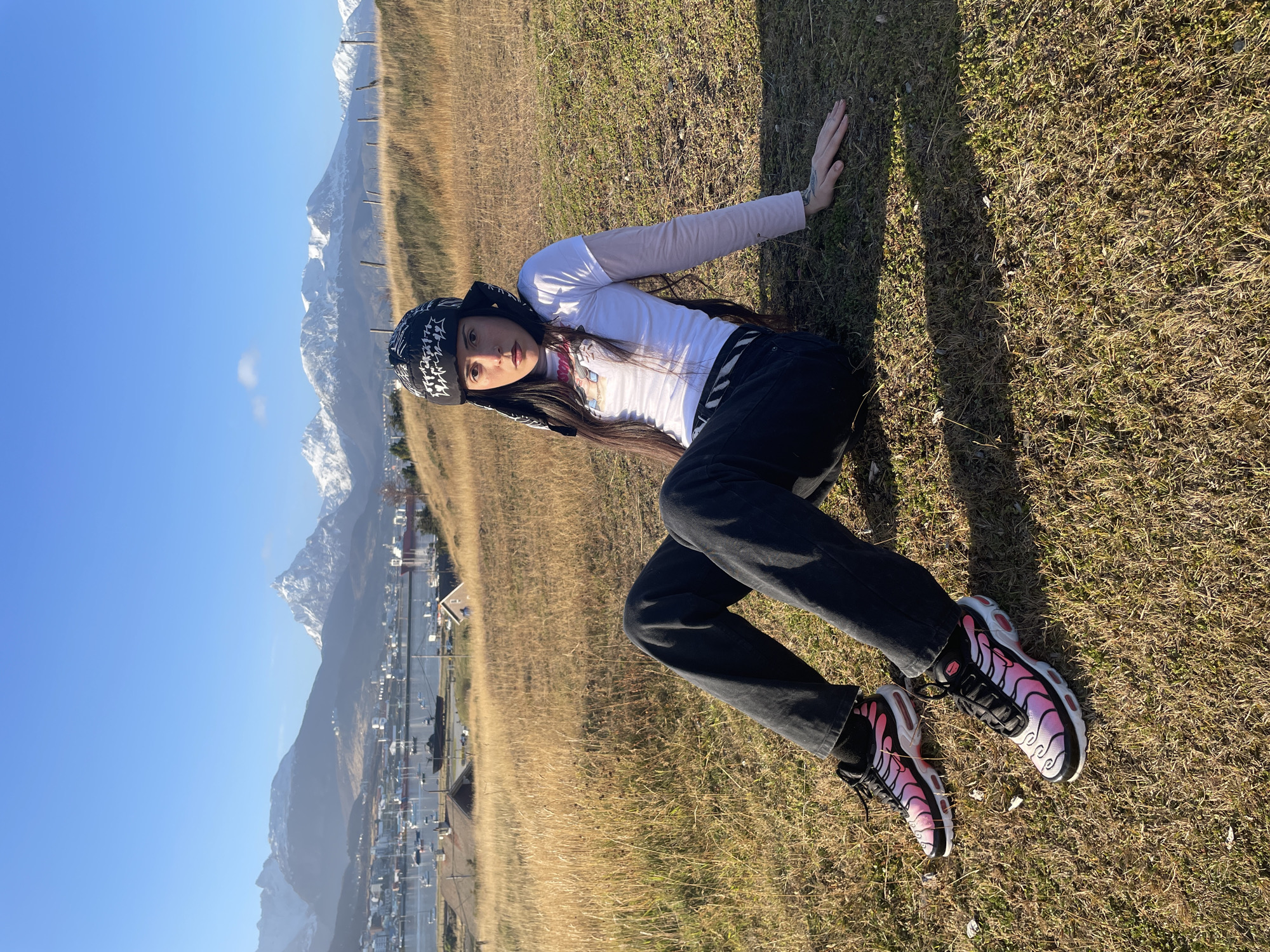 A young woman is lying on grassy ground in Ushuaia, Argentina. She is wearing a black and white bandana, a white long-sleeved shirt, black pants, and colorful sneakers. She is leaning on one hand on the grass. In the background, there are snow-capped mountains and a view of the city and harbor of Ushuaia. The sky is blue.