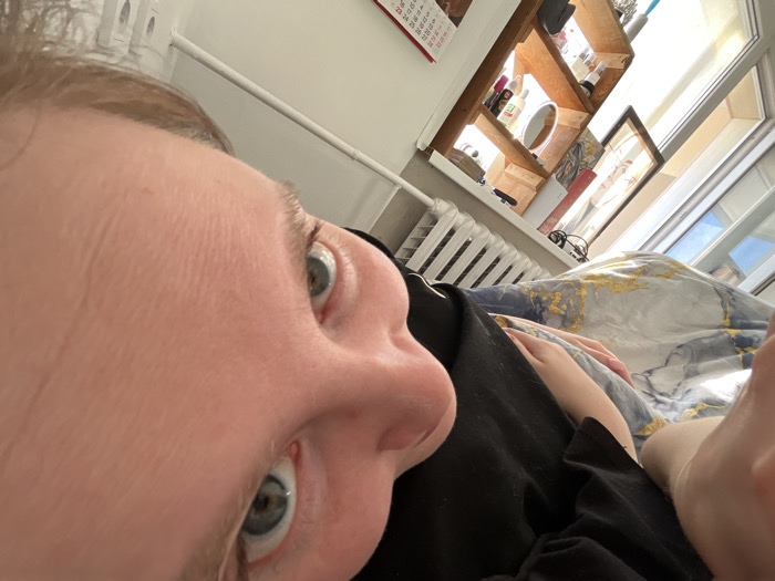 The image displays a low-angle indoor selfie captured from a reclining position. The subject, an individual with fair skin, light-colored hair, and blue/gray eyes, gazes directly at the camera. Visible features include the forehead with fine lines, the bridge of the nose, and both eyes. The individual wears a black garment, and a hand rests on this garment. A bare foot is partially visible in the bottom right. The subject is partially covered by a duvet featuring a white/light gray background with abstract blue and gold/yellow marbled patterns.
The background shows an interior room, likely a bedroom. A bright window in the upper right illuminates the scene, revealing a light blue sky outside. A white radiator with horizontal fins is positioned below the window. Above the radiator, a multi-tiered wooden wall shelf contains various bottles, a small round mirror, and other personal items. A calendar with red text hangs on the wall to the far left. A pair of glasses and a framed object are also visible near the window. The walls are light-colored. The precise location within the city of Chaikovsky, Russia, cannot be determined from the visual information presented.