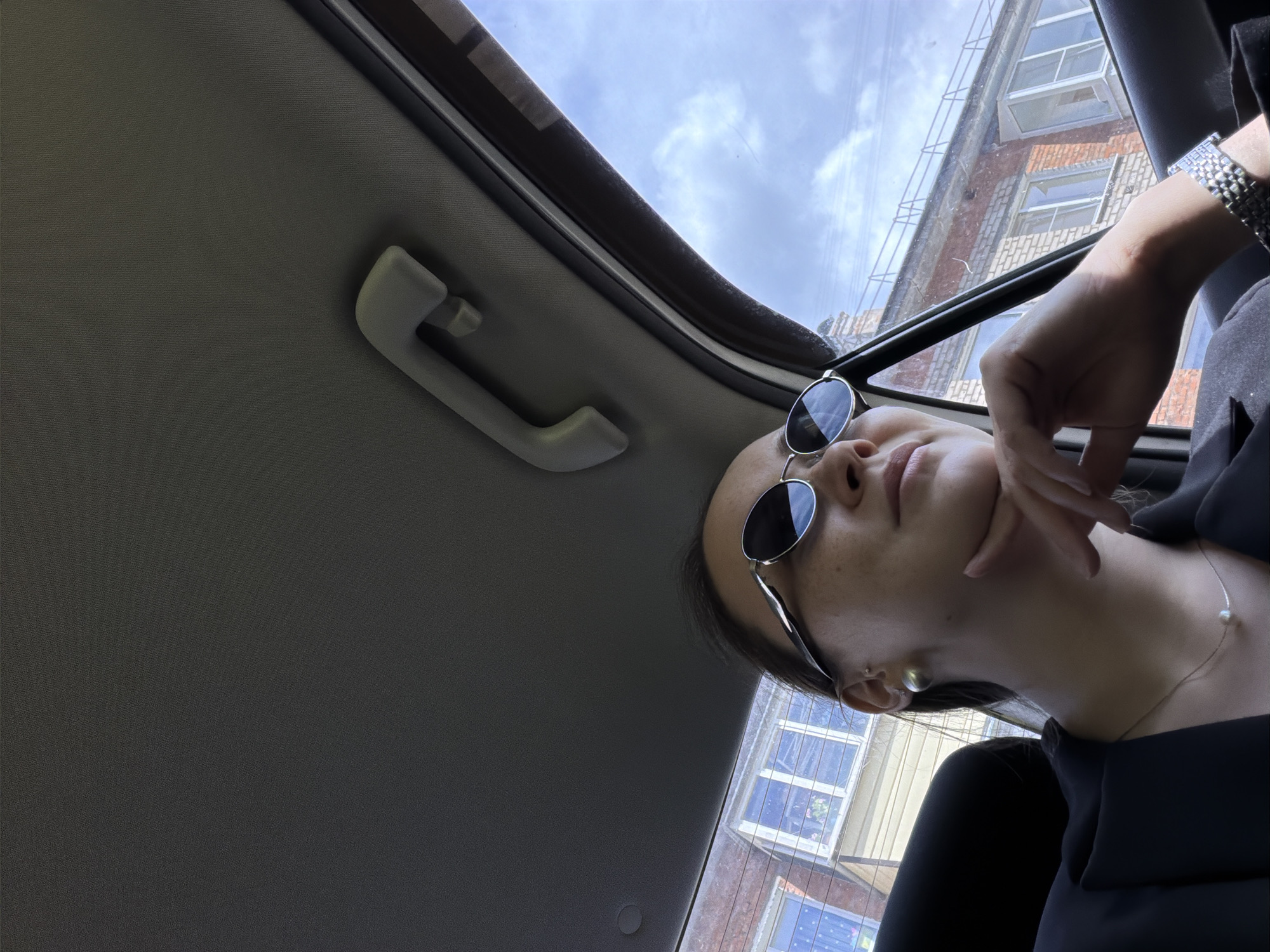 A young woman with dark hair is sitting in the passenger seat of a car. She is wearing sunglasses, a black blazer, a pearl necklace, and a silver watch. She is looking out the window at a brick building with several windows. The sky is visible through the car window, with some clouds. The interior of the car is visible, including the ceiling and a grab handle. The scene appears to be a street in the city of Chaikovsky, Russia, as indicated by the architecture of the building.