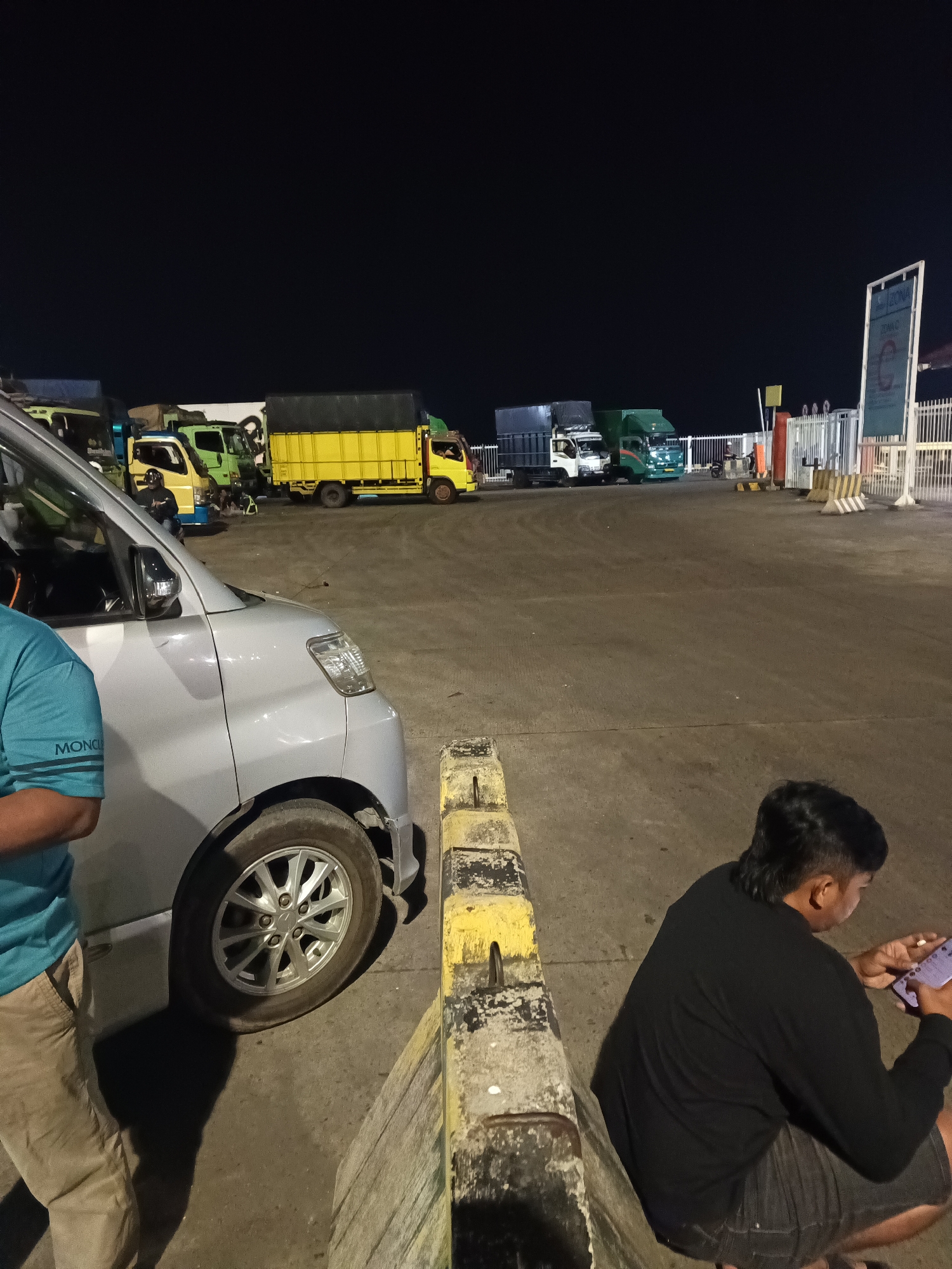 A nighttime scene at the Bakauheni ferry terminal in Indonesia shows several trucks lined up for boarding. A silver minivan is parked in the foreground on the left, partially obscuring a person wearing a blue shirt and tan pants. To the right, a person in a black shirt and shorts is crouched, looking at a smartphone. In the background, a row of trucks, including a prominent yellow truck with a dark tarp covering its load, are parked on the tarmac. A white gate and a sign with green lettering are visible on the far right, indicating the terminal's operational area. The ground is paved, and the lighting suggests it is an outdoor, paved area at night.