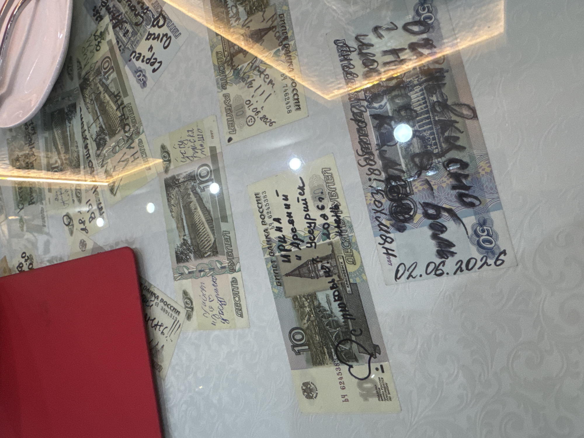 The image is an overhead shot of a glass table with several Russian banknotes laid out on it. Some of the banknotes have handwritten messages and drawings on them. The banknotes appear to be 10 and 50 ruble denominations. One banknote has the date "02.06.2026" written on it. The context of Suifenhe, China, is not evident from the visual information presented in the image. The scene is indoors, with a white textured tablecloth underneath the glass table. A red object, possibly a folder or book, is partially visible in the upper left corner. The lighting suggests a well-lit indoor environment.