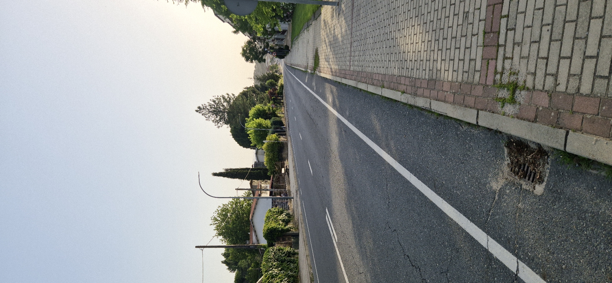 The image shows a street scene in an urban environment. A paved road with a white lane divider runs through the center of the frame, extending into the distance. To the left of the road, there are houses with white walls and red-tiled roofs, surrounded by dense green hedges and trees. A wooden bench is visible in front of one of the houses. To the right of the road, a brick-paved sidewalk is present, bordered by a strip of grass and more greenery. The sky above is clear and light blue, indicating daylight. The overall impression is a quiet, residential street.