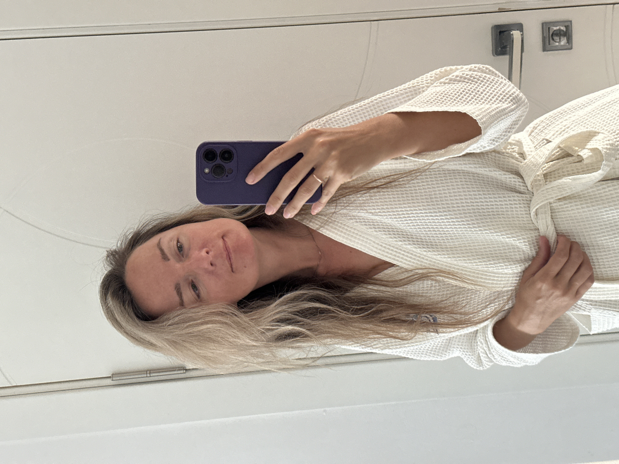 A woman with blonde hair tied in a robe stands in front of a white door and takes a selfie with her phone. She is wearing a white bathrobe tied at the waist. The door has a silver handle and two small silver squares. The floor is not visible. The setting is likely a bathroom or bedroom.