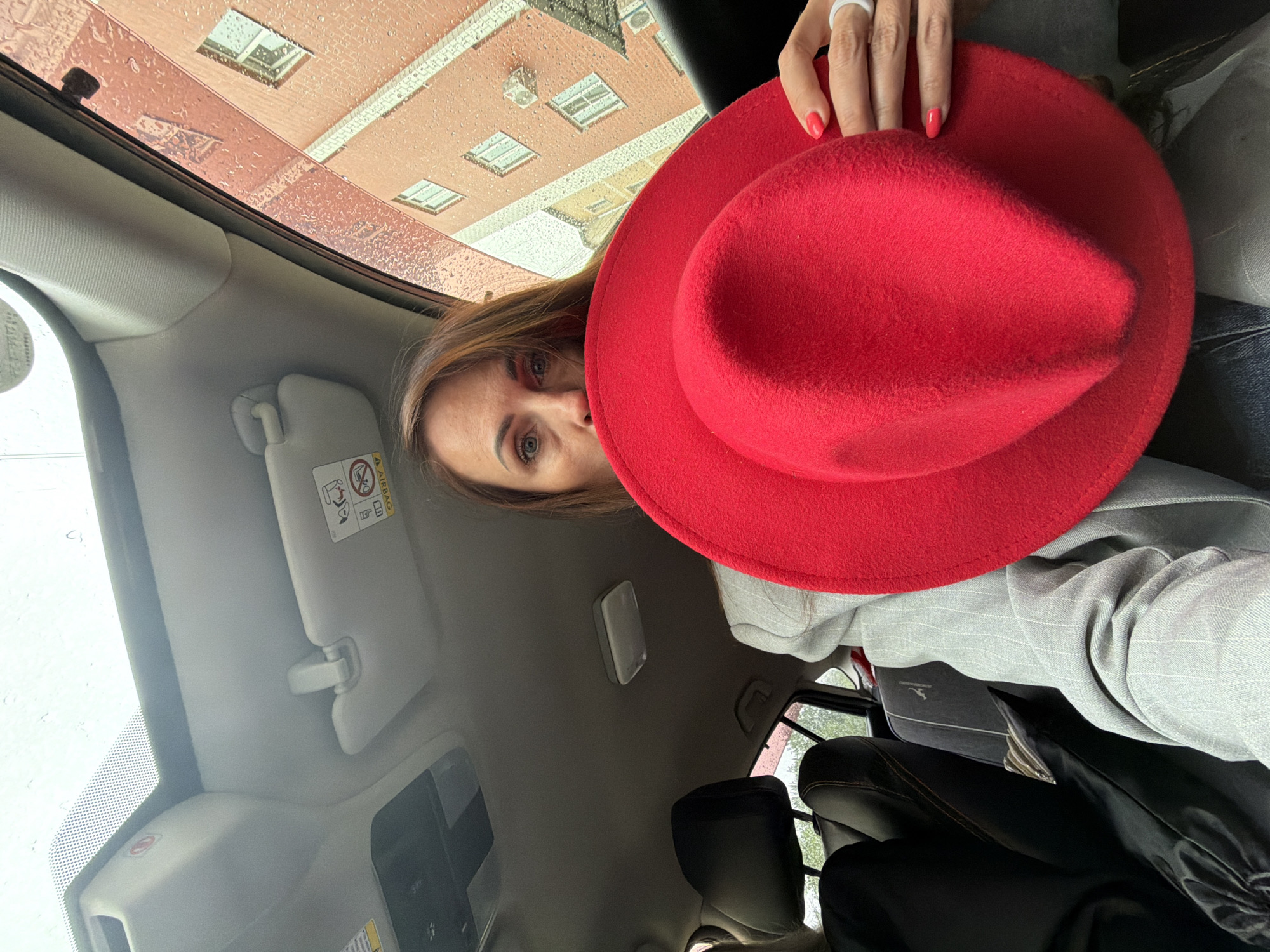 The vertically oriented image presents a tilted perspective from inside a car, capturing a woman partially obscured by a bright red felt fedora-style hat she is holding. Her right eye, nose, and mouth are visible as she looks towards the viewer, revealing light brown hair and a neutral expression. Her left hand, featuring red nail polish and a white ring on the ring finger, holds the hat.
The car's interior is prominent, with a beige headliner and an overhead light. A sun visor is lowered, displaying visible warning labels, including the word "AIRBAG" and a graphic symbol cautioning against using a rear-facing child seat in the front with an active airbag. Parts of black car seats and dashboard elements are also visible.
Through the car's window, which is covered in raindrops, an exterior scene unfolds. A red-brown brick building with multiple square windows is visible, reflecting some light. The presence of raindrops indicates rainy weather. The overall lighting suggests the image was taken during the daytime. The location context for this scene is Birobidzhan, Russia.