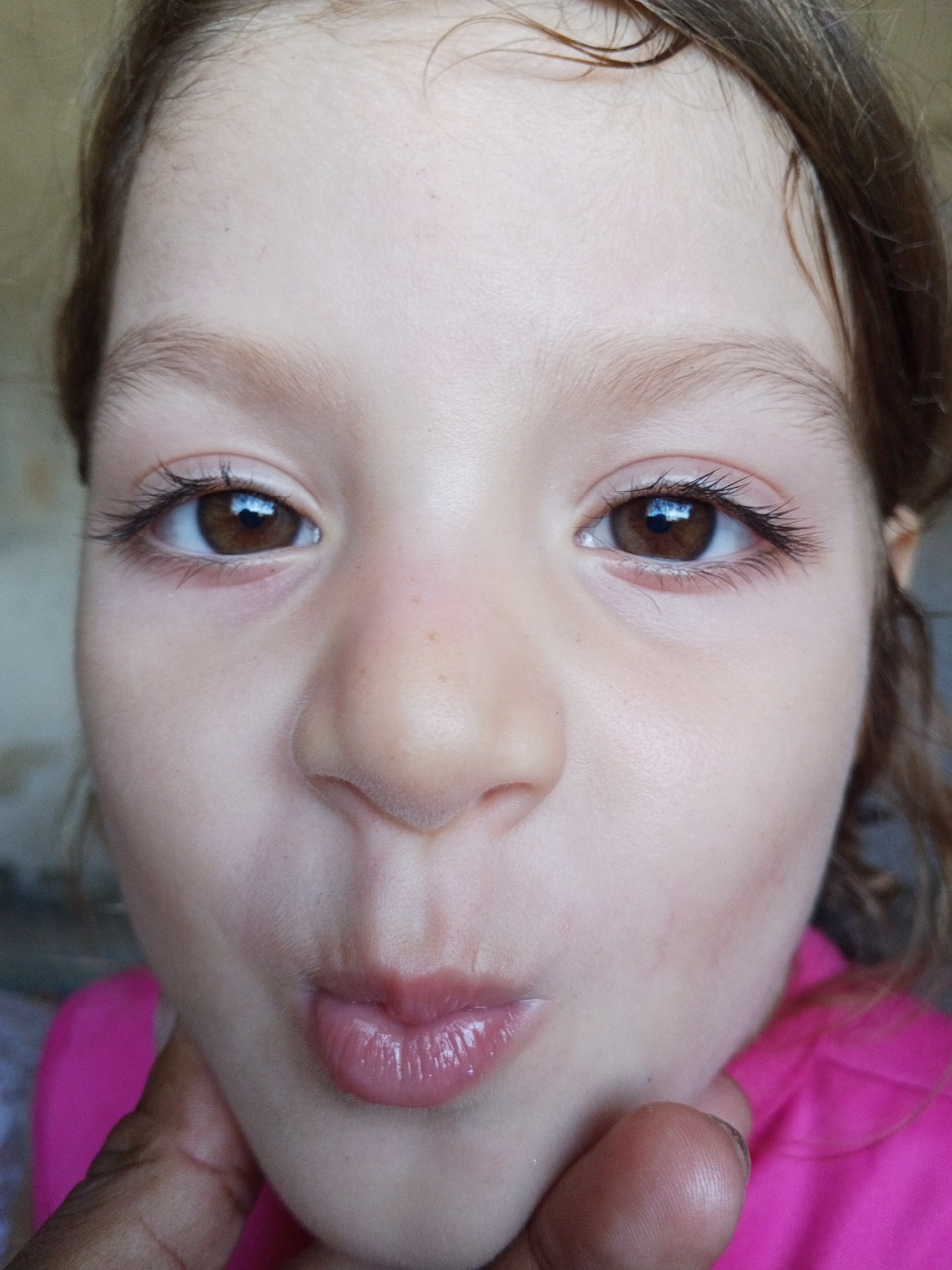 The image is a close-up portrait of a young girl's face. Her eyes are wide open and brown, with long eyelashes. Her nose is small and upturned, and her lips are slightly parted, appearing glossy. She has fair skin with a few faint freckles. Her eyebrows are light brown and thin. Her hair is brown and appears to be tied back or falling loosely around her face. Her head is tilted slightly to the left. A finger, likely belonging to an adult, is visible near her mouth, supporting her chin. The background is out of focus and appears to be a light-colored wall. The overall lighting suggests an indoor environment.