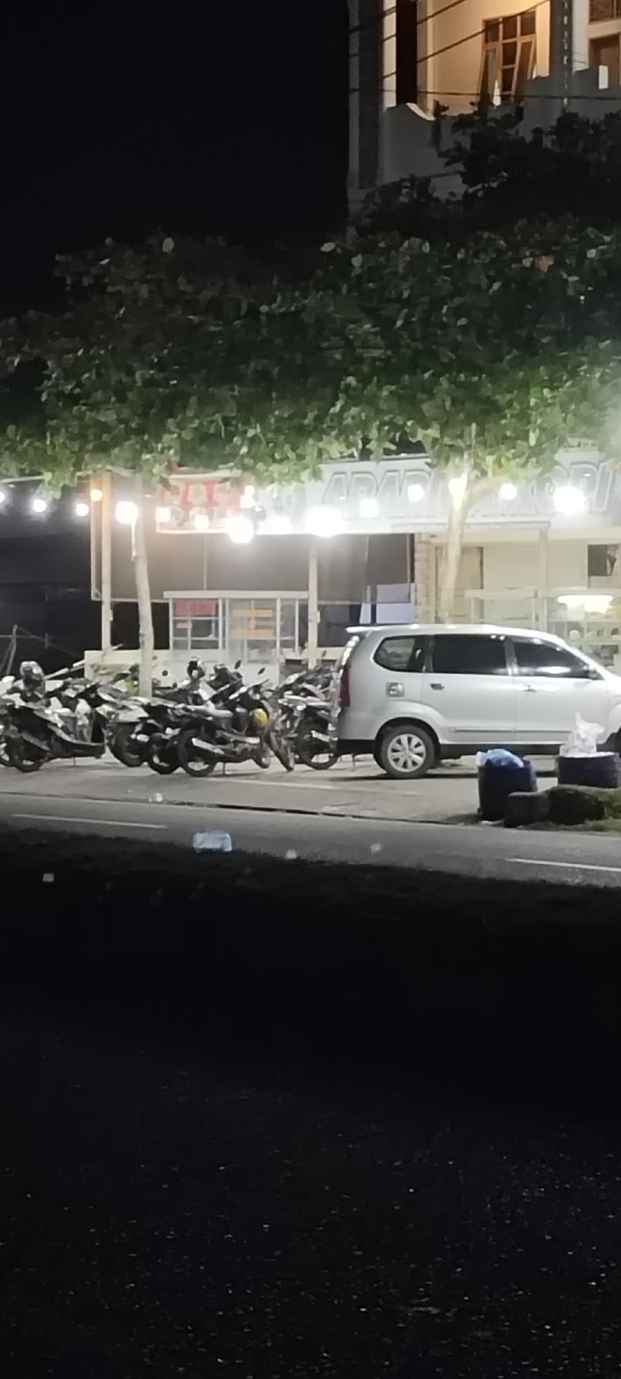 The image depicts a street scene at night in Unknown, Unknown. In the background, a brightly lit building with a sign that appears to read "APABARI" can be seen. A row of motorcycles is parked in front of the building, with a silver van parked at the end of the row. Trees hang overhead. The foreground shows the road, which is dark.