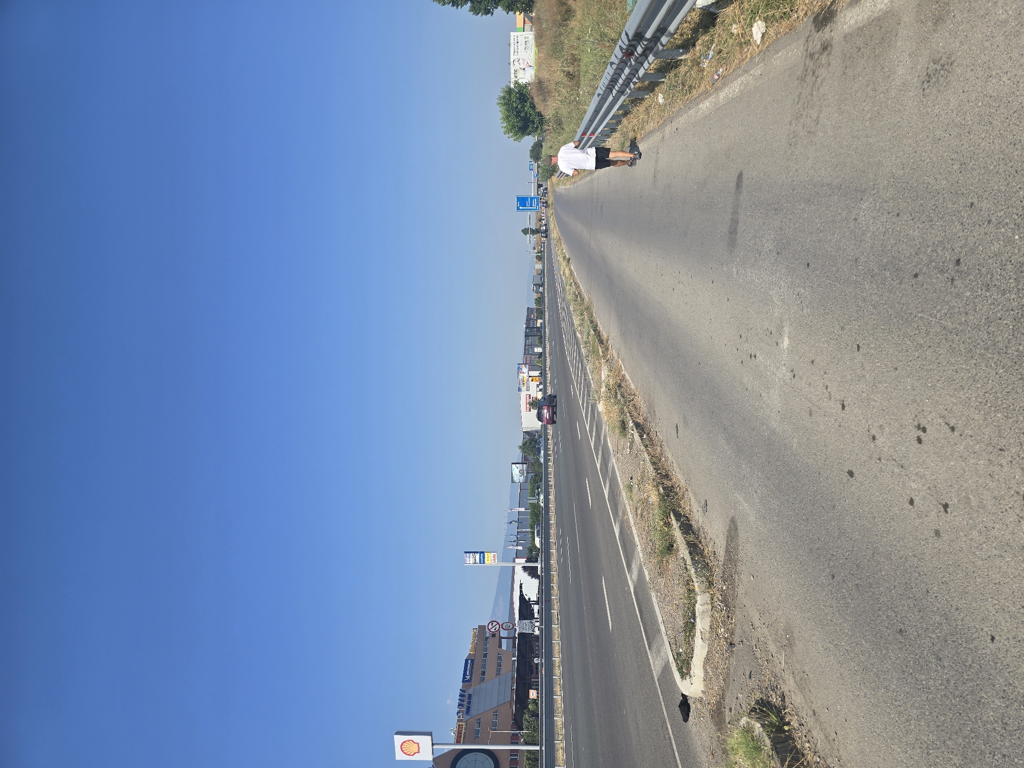 This image depicts a bright, clear day with a vast expanse of blue sky dominating the upper portion. In the foreground, a two-lane road stretches into the distance, with the right lane being used by a lone individual jogging away from the camera. The jogger is wearing a white t-shirt and black shorts. The road surface shows signs of wear, with some dark stains and patches of dry grass and debris lining the edges, particularly along the guardrail on the right.
The left side of the road features a median with more dry vegetation. Beyond the median, an elevated highway or a similar road is visible, with cars and trucks in transit. Prominently featured on the left is a Shell gas station sign and a building with a circular clock face. Further down the road on the left, several commercial buildings and billboards are visible.
On the right side of the image, beyond the road and guardrail, is a dense patch of dry grass and vegetation, with some trees in the background. A large billboard with an advertisement is also visible on the right. The overall setting appears to be an industrial or commercial area on the outskirts of a city, likely near a major road or highway. Based on the signage and landscape, this location is the A1 motorway (Trakia Highway) near the western entrance to Sofia, Bulgaria, possibly in the vicinity of the "Orion" commercial zone or near the business park at Tsaritsa Yoanna Boulevard.