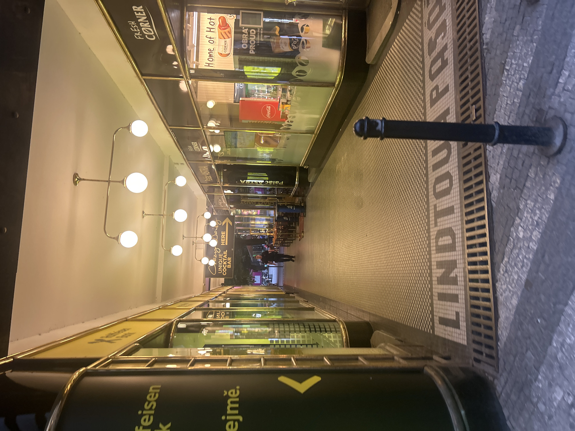 This medium-shot image captures a well-lit indoor shopping or pedestrian passage, identified by the floor inscription "LINDTOU PASA" (Lindtova Pasáž), located in Prague, Czechia. The passage is long and relatively narrow, flanked on both sides by glass storefronts displaying various goods and advertisements.
On the left, a series of modern, globe-shaped pendant lights illuminate the white ceiling and the passage. The storefronts on both sides feature gold-colored trim and reflections of the opposite side. Visible signs include "FRESH CORNER" multiple times, "Home of Hot," "OBRA PROUD," and a "Coca-Cola" display. Further down the passage on the left, a sign reads "Palác ASTRA," likely indicating the building's name. On the right, towards the end of the visible section, a prominent sign for "CRAZY DAISY UNIQUE COCKTAIL BAR" with an arrow pointing further in suggests an establishment. Partially visible text near the bottom left includes "feisen k ejmě.", likely related to Raiffeisen Bank.
The floor of the passage is covered in distinct tile patterns: a dark checkerboard pattern runs down the center, while a lighter, more intricate mosaic pattern spelling out "LINDTOU PASA" occupies a central strip. On the right side, the tiled floor transitions to a grid grate and then to an outdoor cobblestone path, where a black bollard stands. A few indistinct figures of people are visible in the distance, walking or standing in the passage. The artificial lighting suggests it is either evening or an interior space designed with consistent illumination.