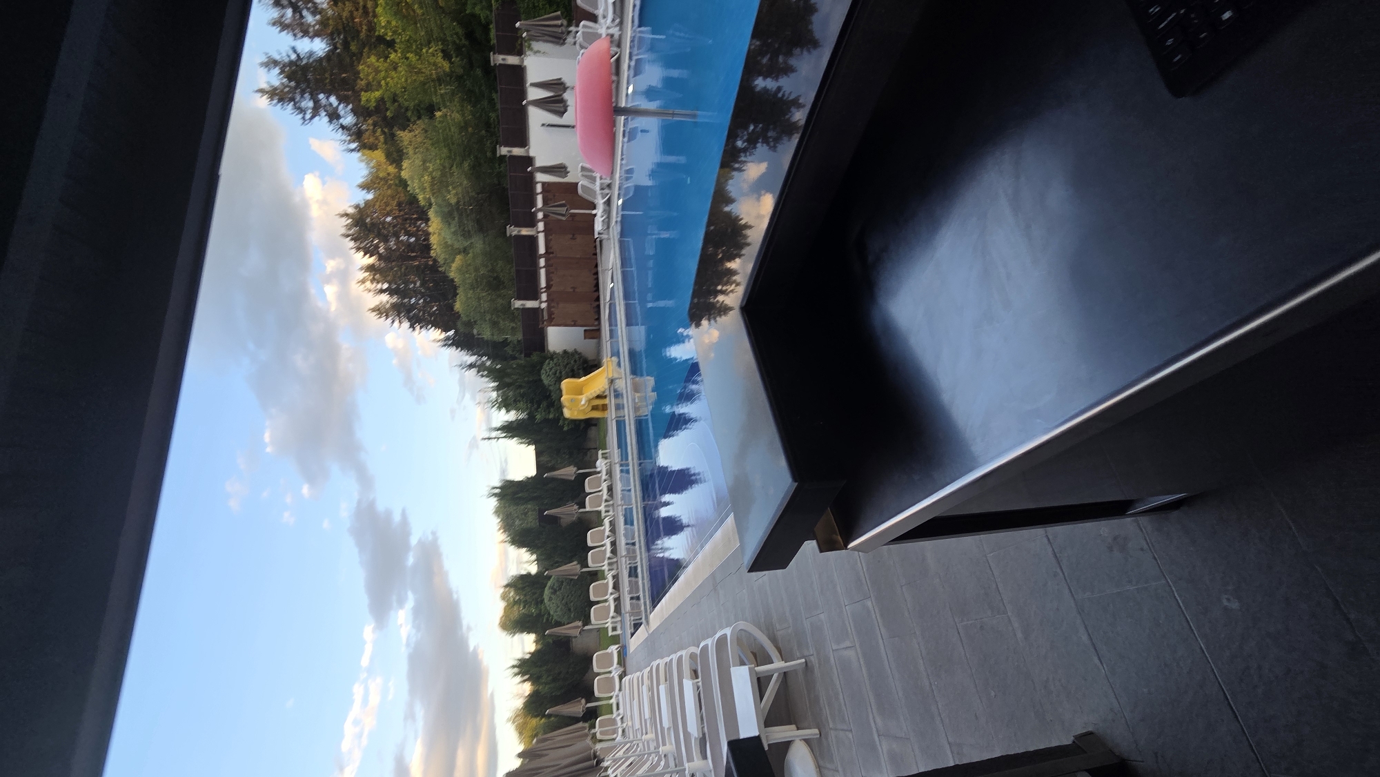The image shows an outdoor swimming pool area at dusk. The pool is blue and reflects the sky, which is a mixture of blue with scattered clouds. A yellow water slide is visible at one end of the pool. Around the pool are rows of lounge chairs, some with umbrellas. In the foreground, a dark, reflective counter or table is visible, with a keyboard on its surface. The area is surrounded by trees and a fence. The scene appears to be in a resort or a private recreational area.