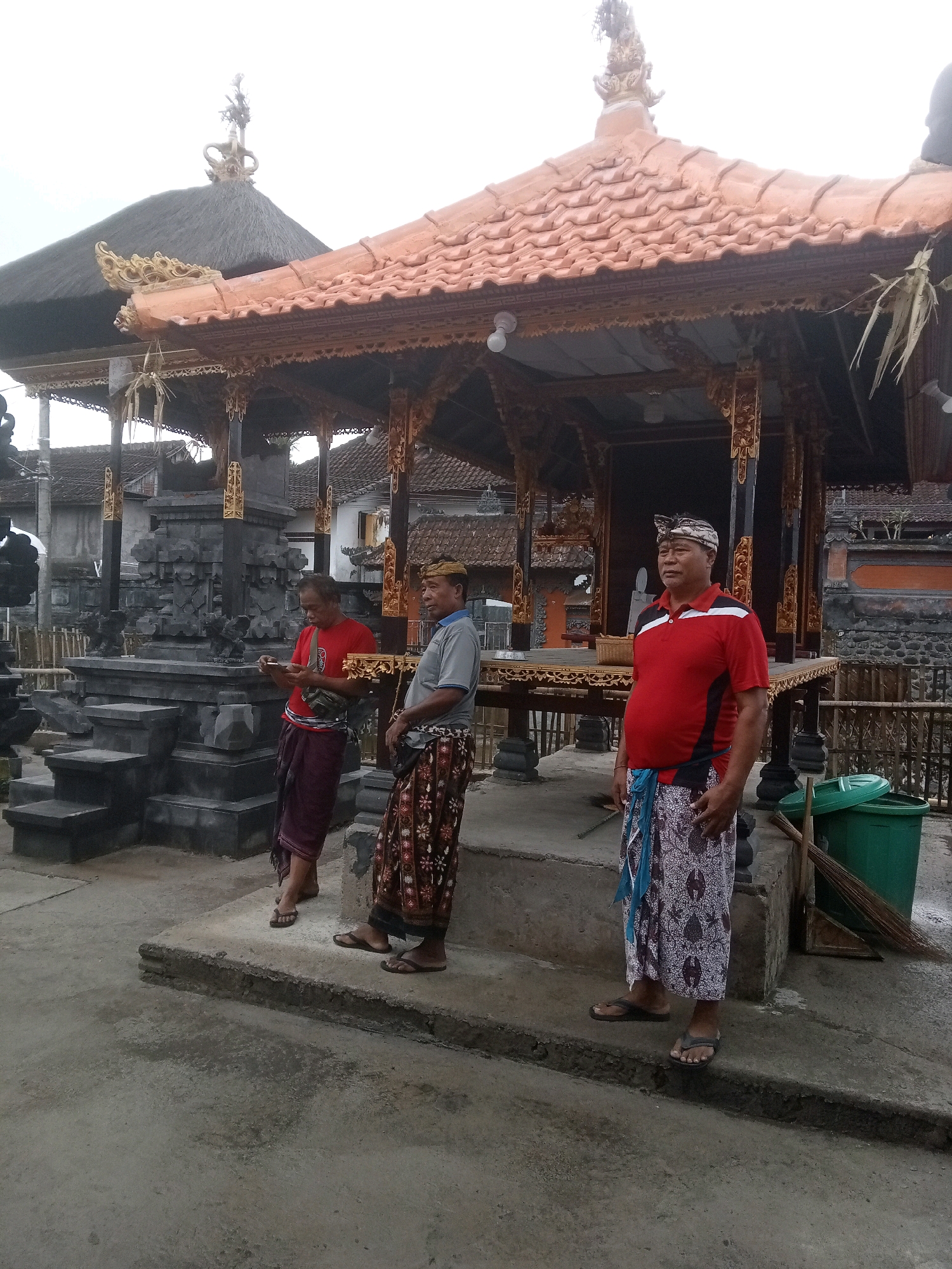 The image depicts three adult males standing outside a structure with a tiled, orange roof and elaborate wooden carvings in what appears to be a temple complex in Mendoyo, Indonesia. The men are dressed in traditional Indonesian attire, including sarongs. The man on the far left wears a red t-shirt and a maroon sarong. The middle man wears a grey polo shirt and a patterned sarong. The man on the right wears a red and white polo shirt, a patterned sarong, and a traditional headpiece. Behind the men is a tiered stone structure and the aforementioned temple building. In the background, there are other buildings with tiled roofs and stone walls. To the right of the main structure, there is a green trash can and a broom. The ground is paved with concrete. The sky is overcast.