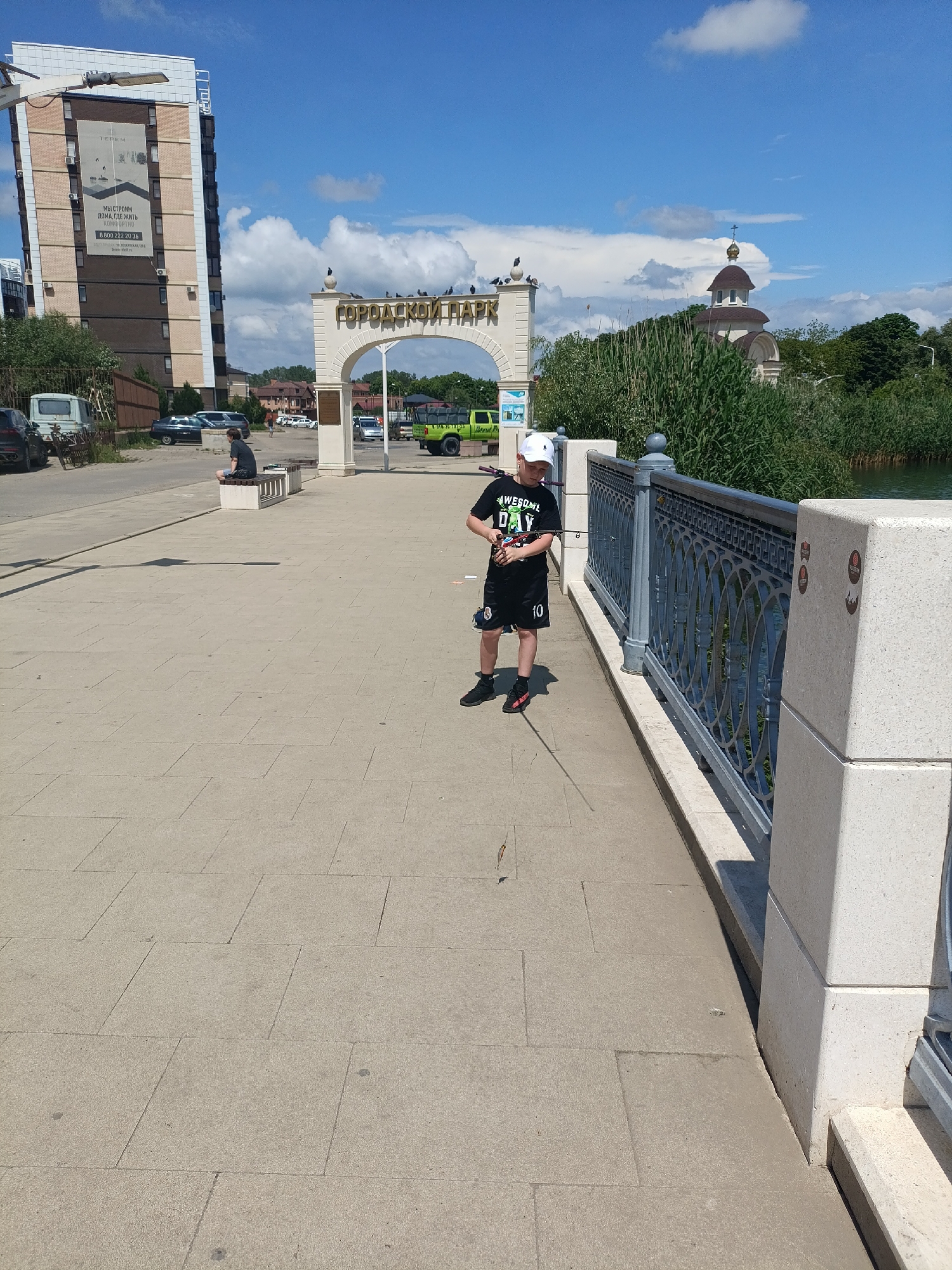 The image depicts a young boy fishing on a bridge in Goryachiy Klyuch, Russia. He is wearing a white cap, a black t-shirt with "AWESOME DAY" printed on it, black shorts with the number "10" and a small emblem on it, and black and red sneakers. He is holding a fishing rod. The bridge has a grey metal railing with ornamental designs and is paved with grey stone tiles. In the background, there is an arched entrance marked "ГОРОДСКОЙ ПАРК" (City Park), a tall building with advertisements, a person sitting on a bench, and a church with a golden dome. The sky is blue with scattered clouds. A body of water is adjacent to the bridge. Cars are parked on the street behind the bench.