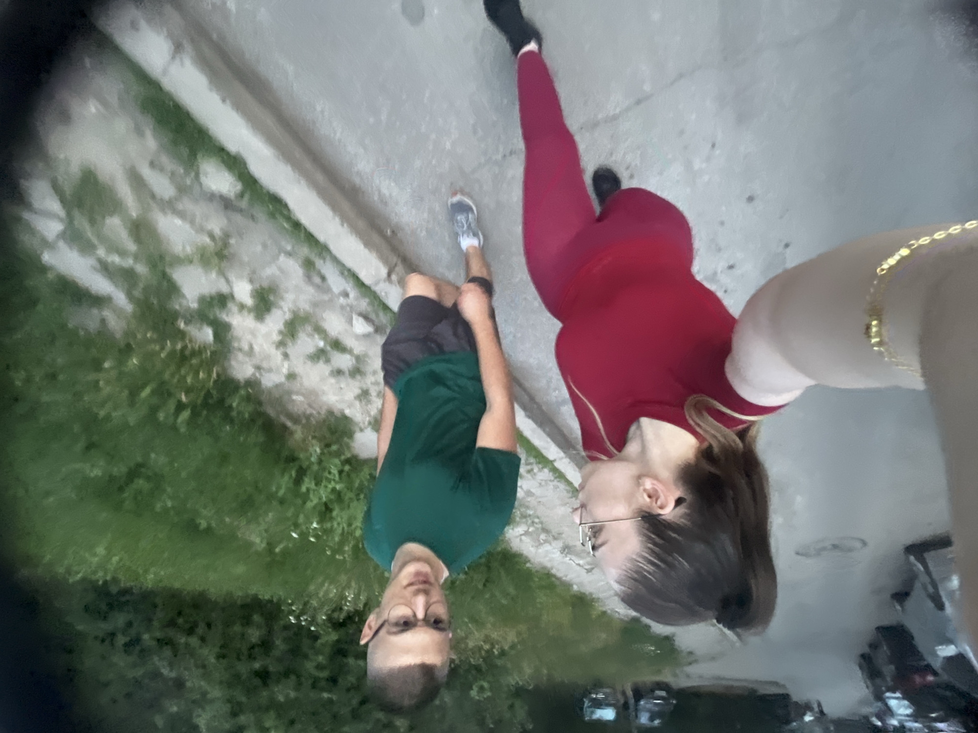 In a street scene, two individuals, a woman and a man, are captured from a low-angle, wide-angle perspective. The woman, in the foreground and to the left, is wearing a red top and matching leggings, with her hair in a high ponytail. She is looking towards the man. The man, to the right and slightly behind her, is wearing a green t-shirt and dark shorts, with glasses. They appear to be walking or standing on a paved surface, possibly a sidewalk or street, with parked cars visible in the background on the left. To the right of the paved area, there is a grassy embankment with some vegetation. The lighting suggests it is either dusk or an overcast day. The overall impression is a casual outdoor setting.